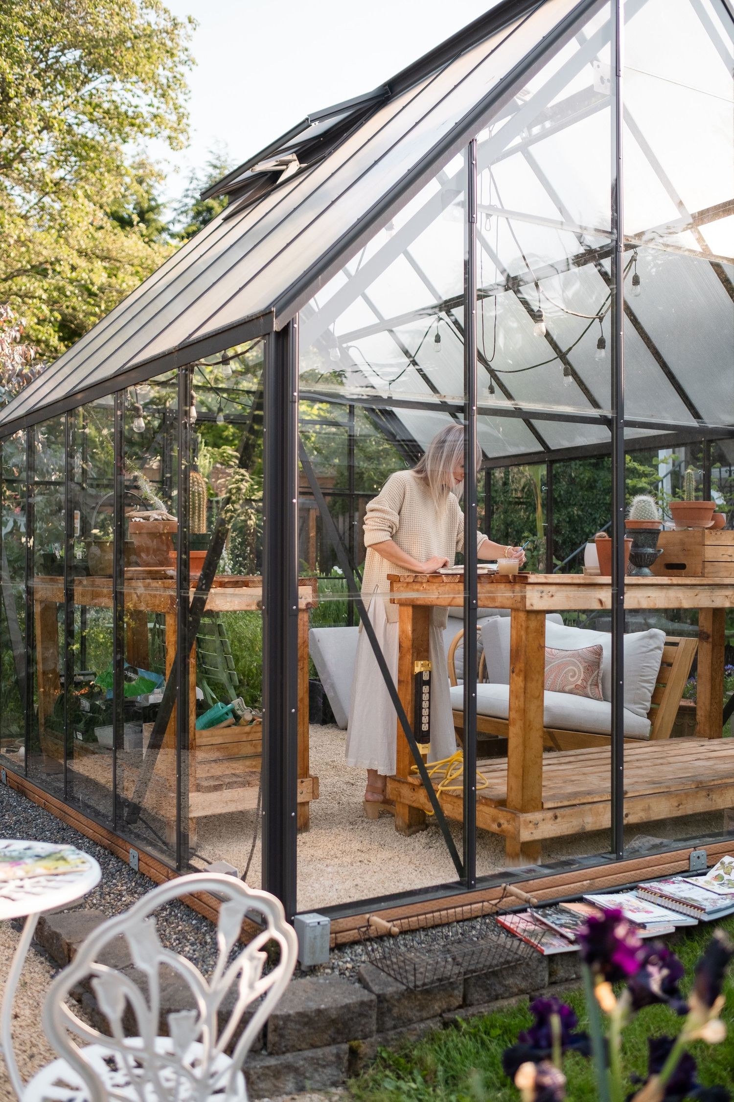 Woman working inside a modern black-framed glass greenhouse surrounded by garden furniture and lush plants at sunset.