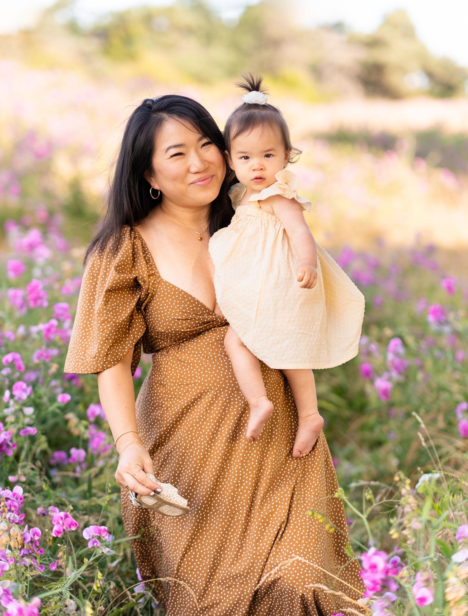 Mother and child in brown dress sit among blooming purple wildflowers during golden hour photo session.