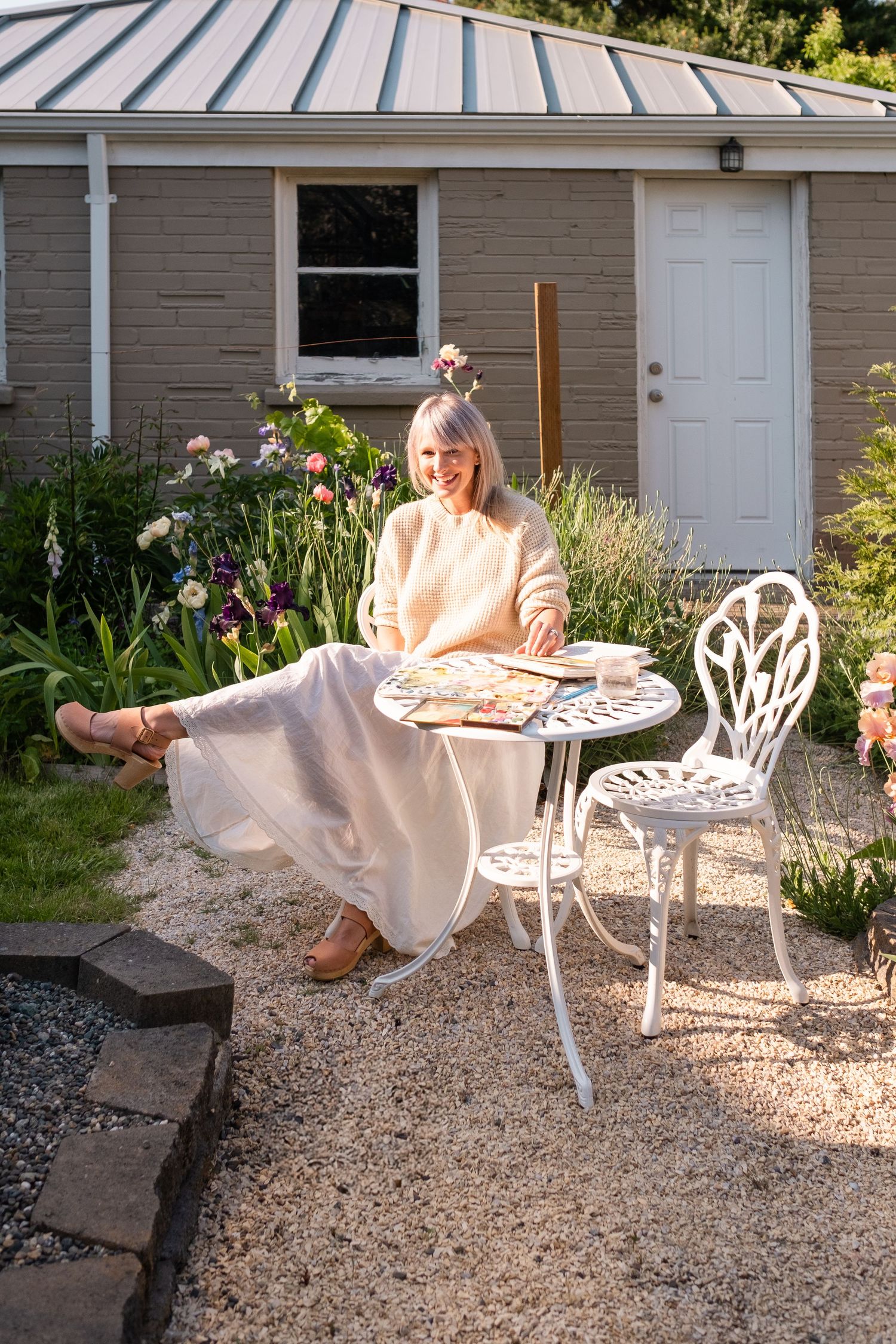 Smiling blonde woman in cream sweater and white skirt sitting at white garden bistro table with watercolor palette outdoors.
