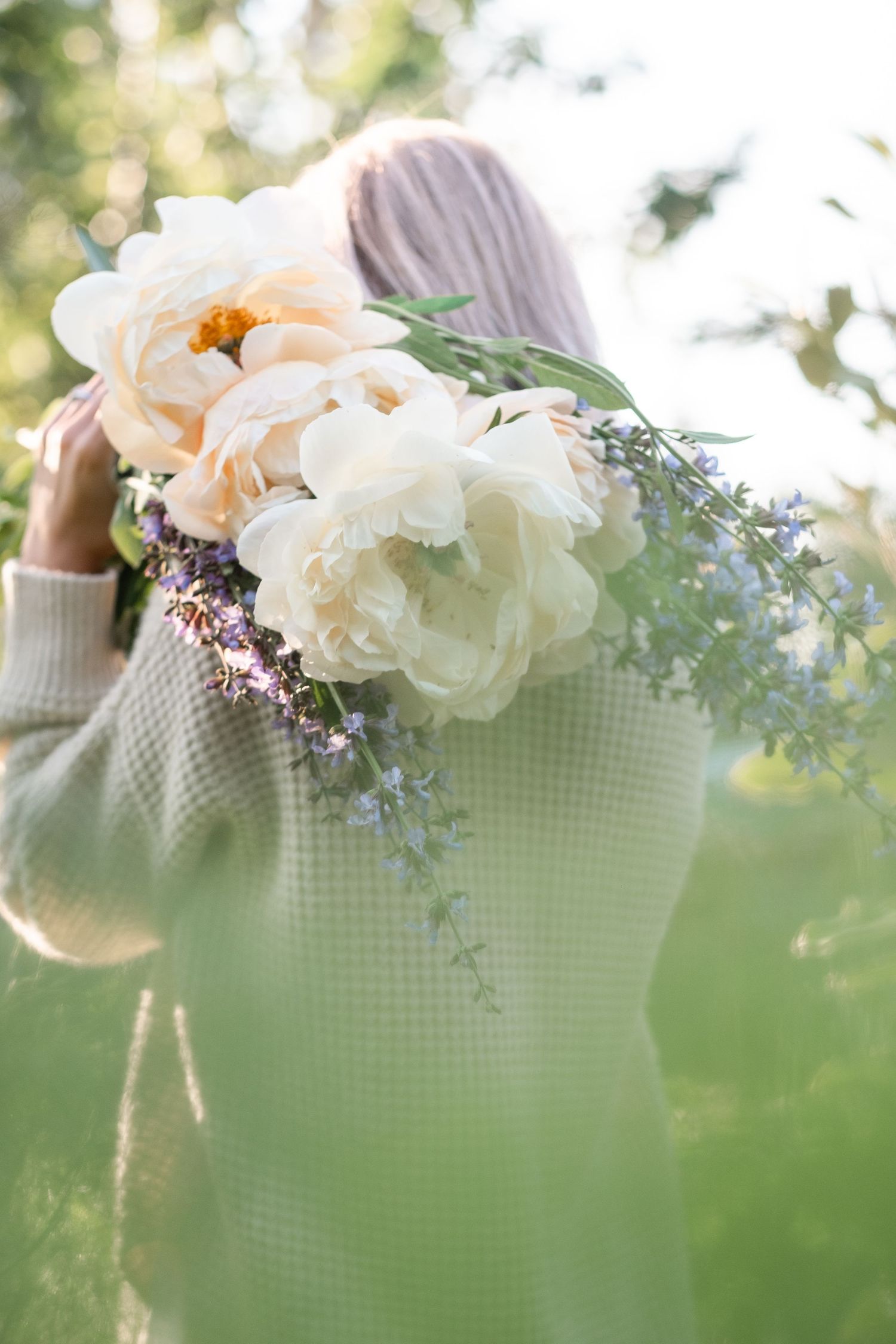 Back view of woman in beige sweater holding white peony bouquet over shoulder in garden.