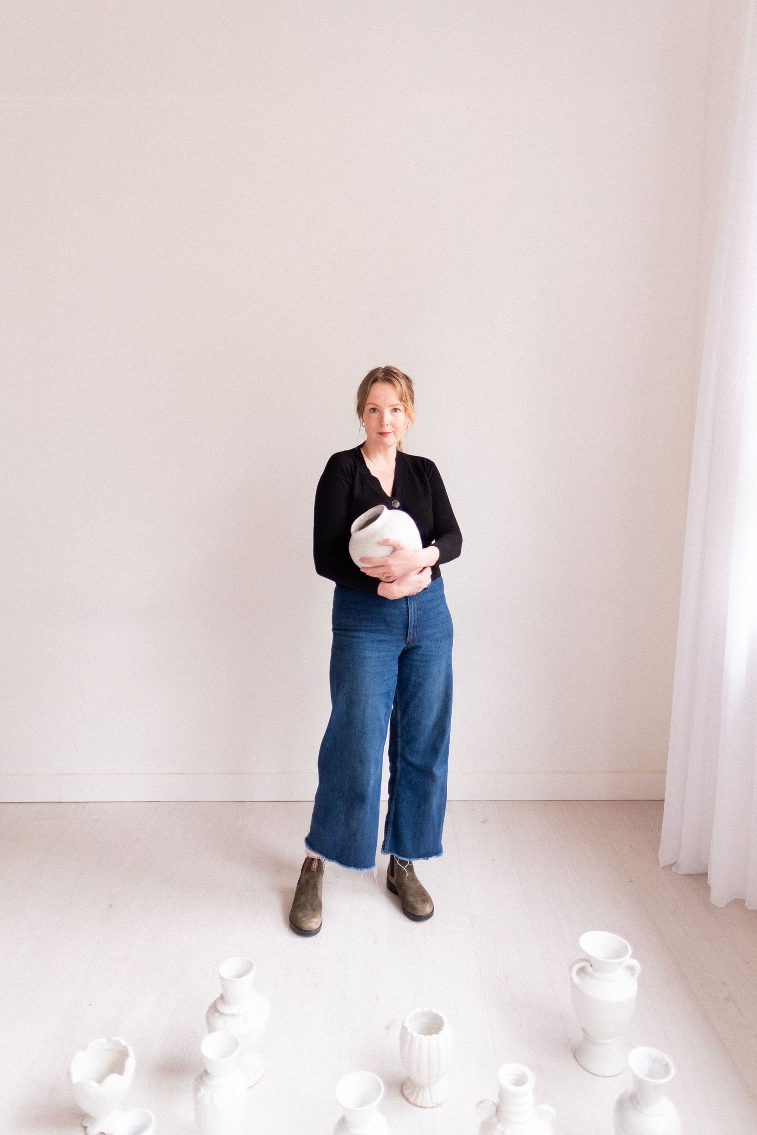 Artist standing in minimalist white studio space surrounded by white ceramic vessels on floor.