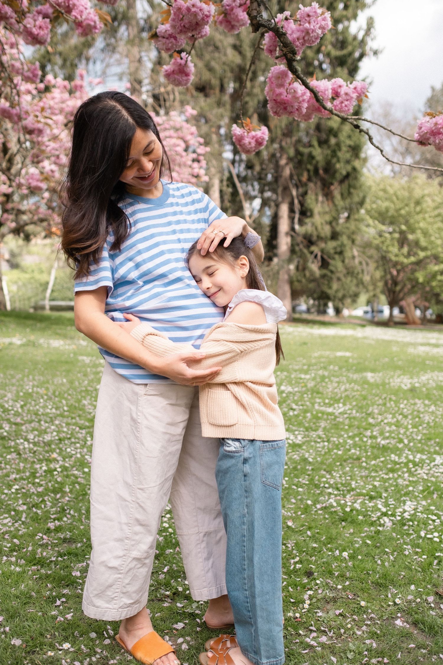 Two people embrace while standing on grass with pink cherry blossoms overhead and white flowers scattered below.
