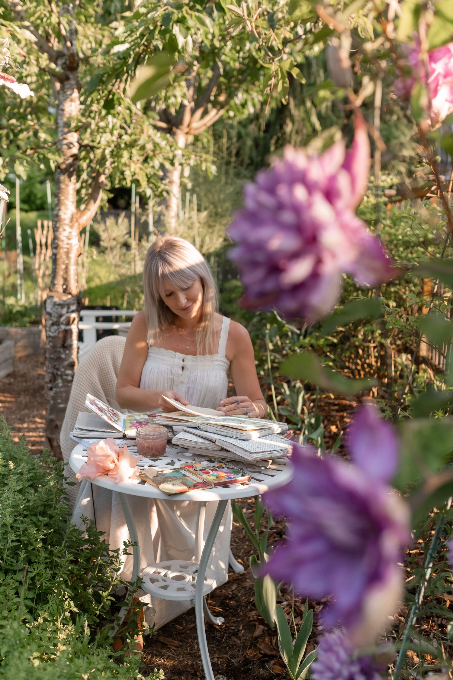 Blonde woman sitting at white garden table painting in sketchbook surrounded by purple flowers.