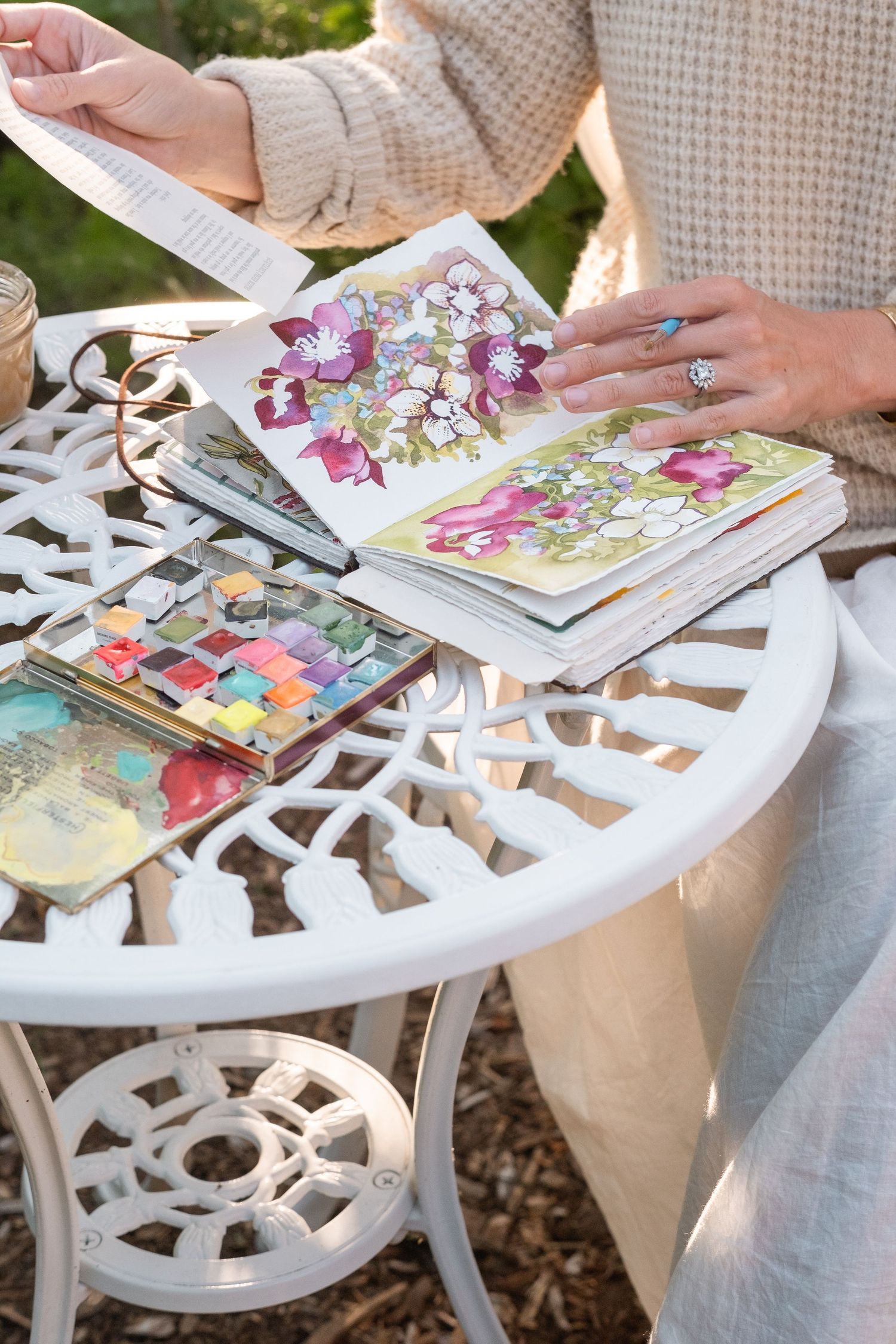 Close-up of watercolor sketchbook with floral paintings on a white ornate garden table.
