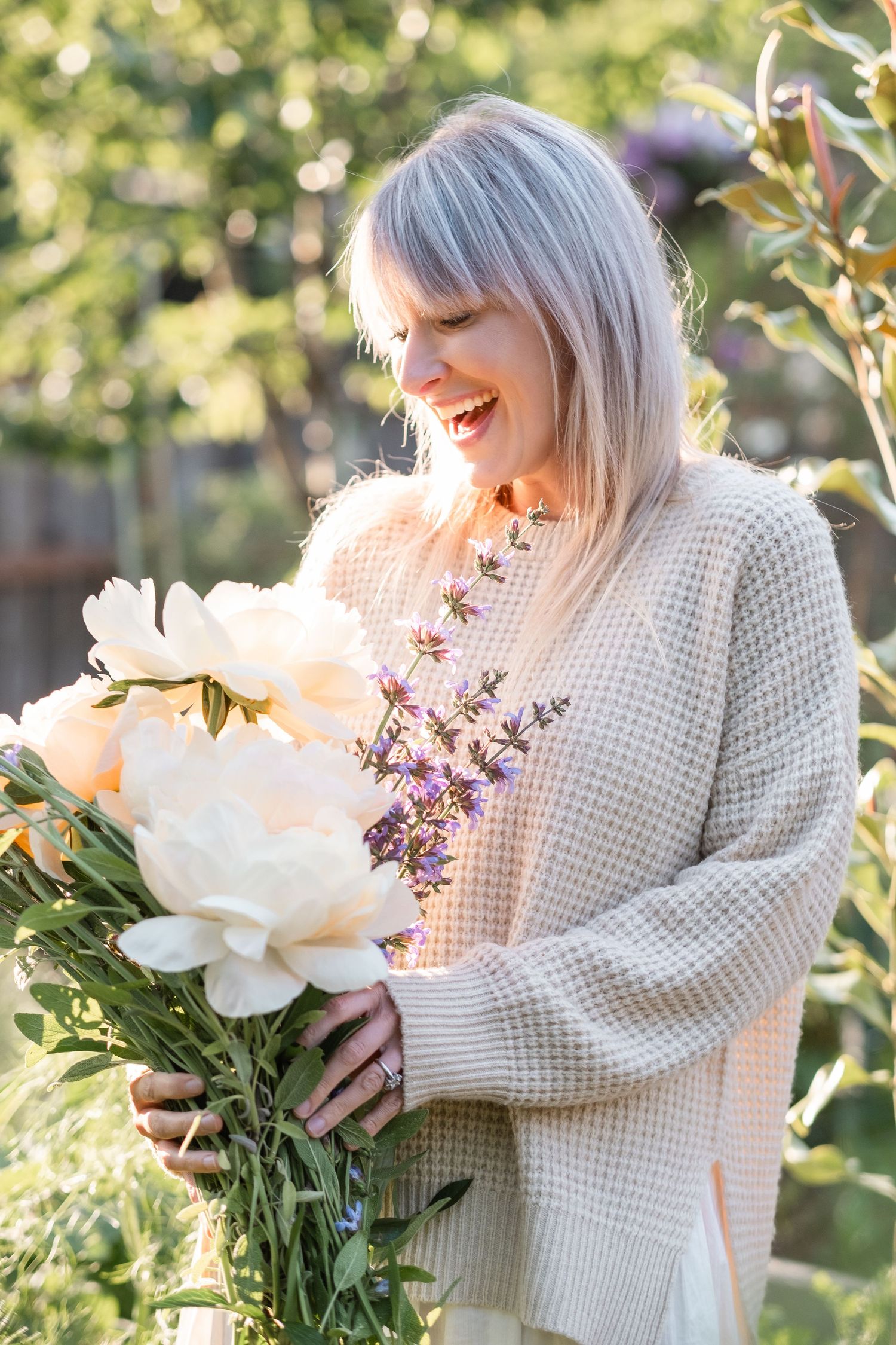 Happy blonde woman in beige sweater holding bouquet of white peonies in sunny garden.
