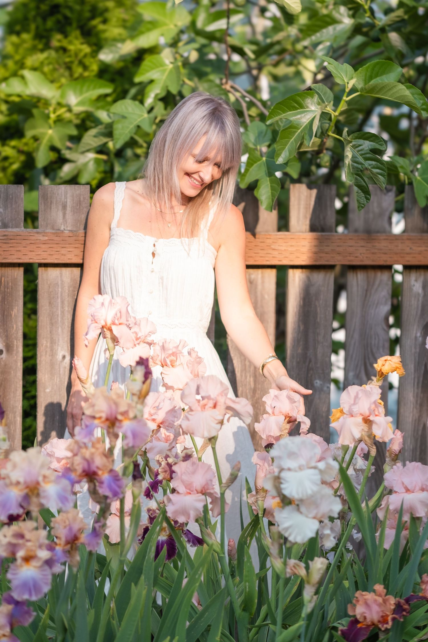 Woman in white sundress reaching toward blooming pink iris flowers by a wooden garden fence.