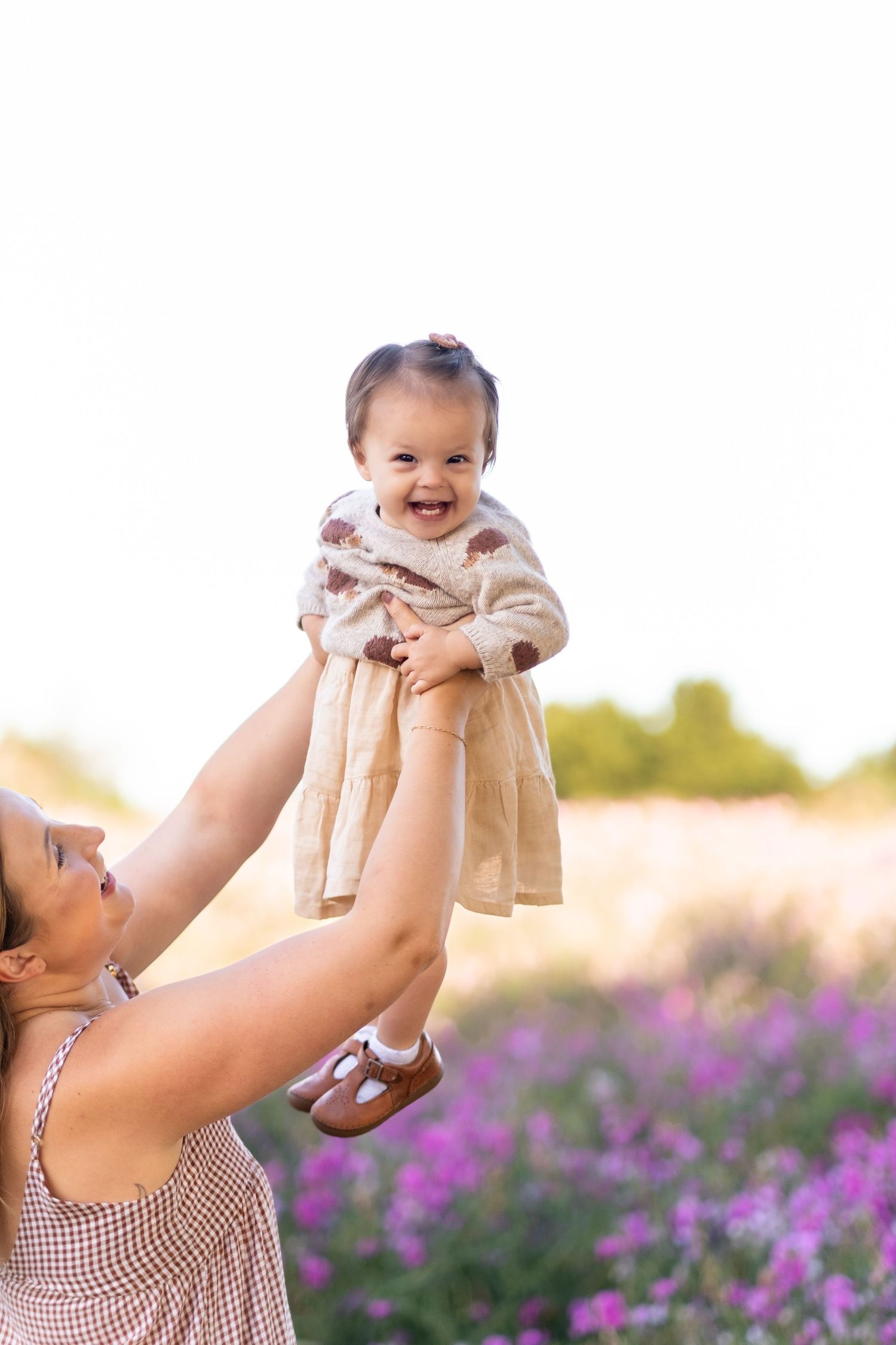A caregiver lifts a joyful baby wearing a tan dress in a blooming purple flower field at sunset.