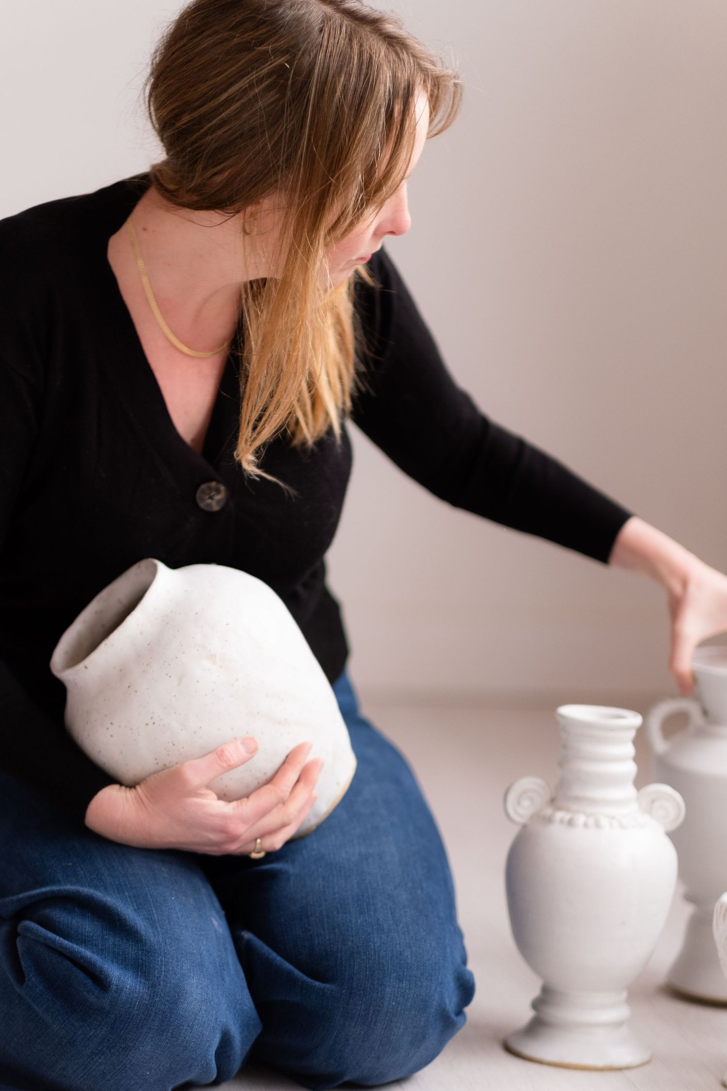 Person in black sweater arranging white ceramic vases and decorative pieces on a light background.