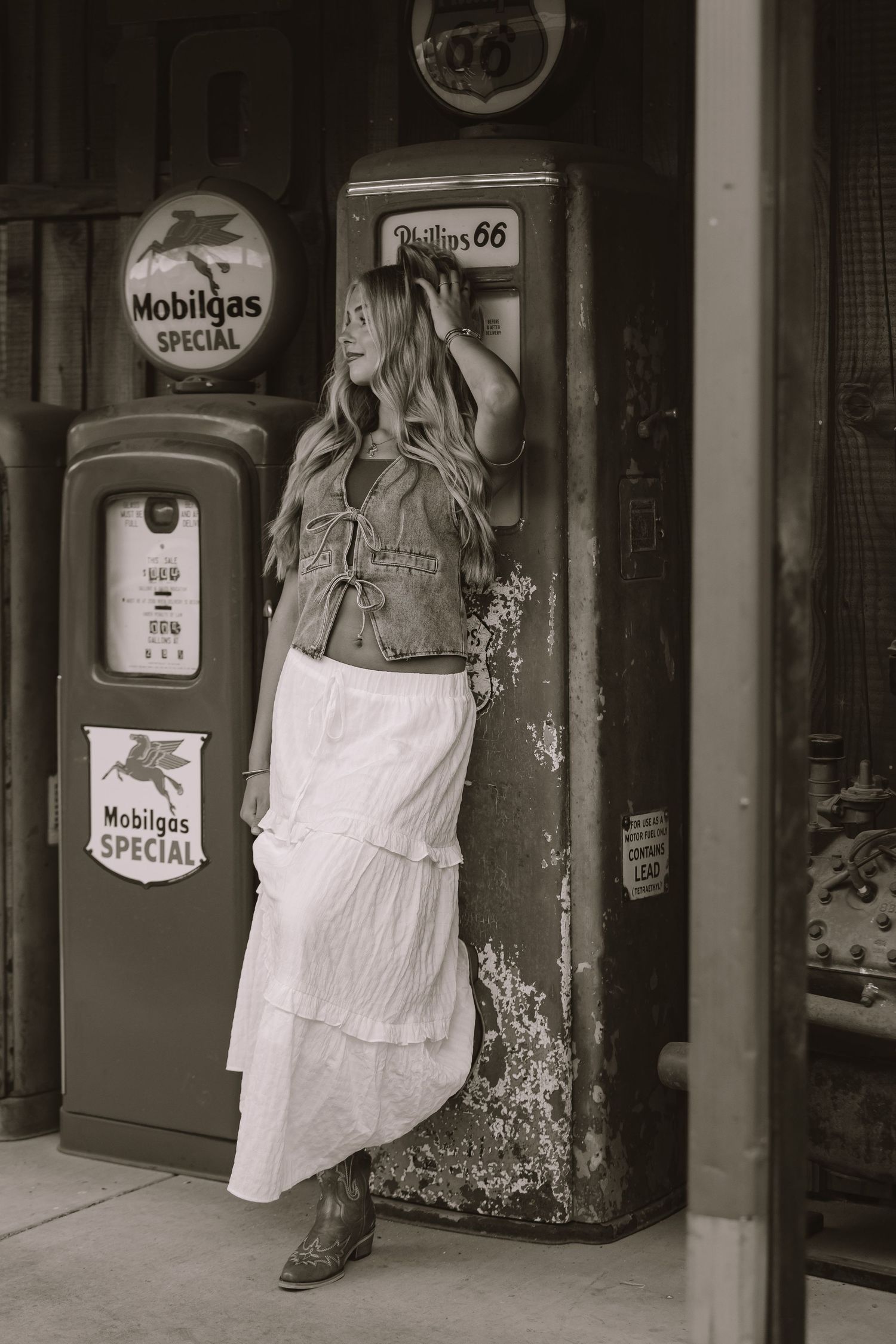 Vintage-style photo of a person in a white dress and denim jacket posing by retro Mobilgas gas pumps.