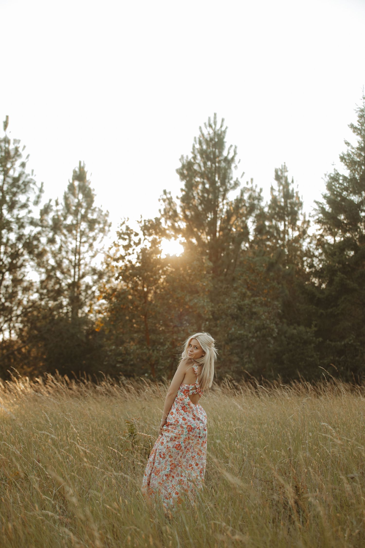 Floral dress twirls through tall grass with pine trees in the background at sunset.