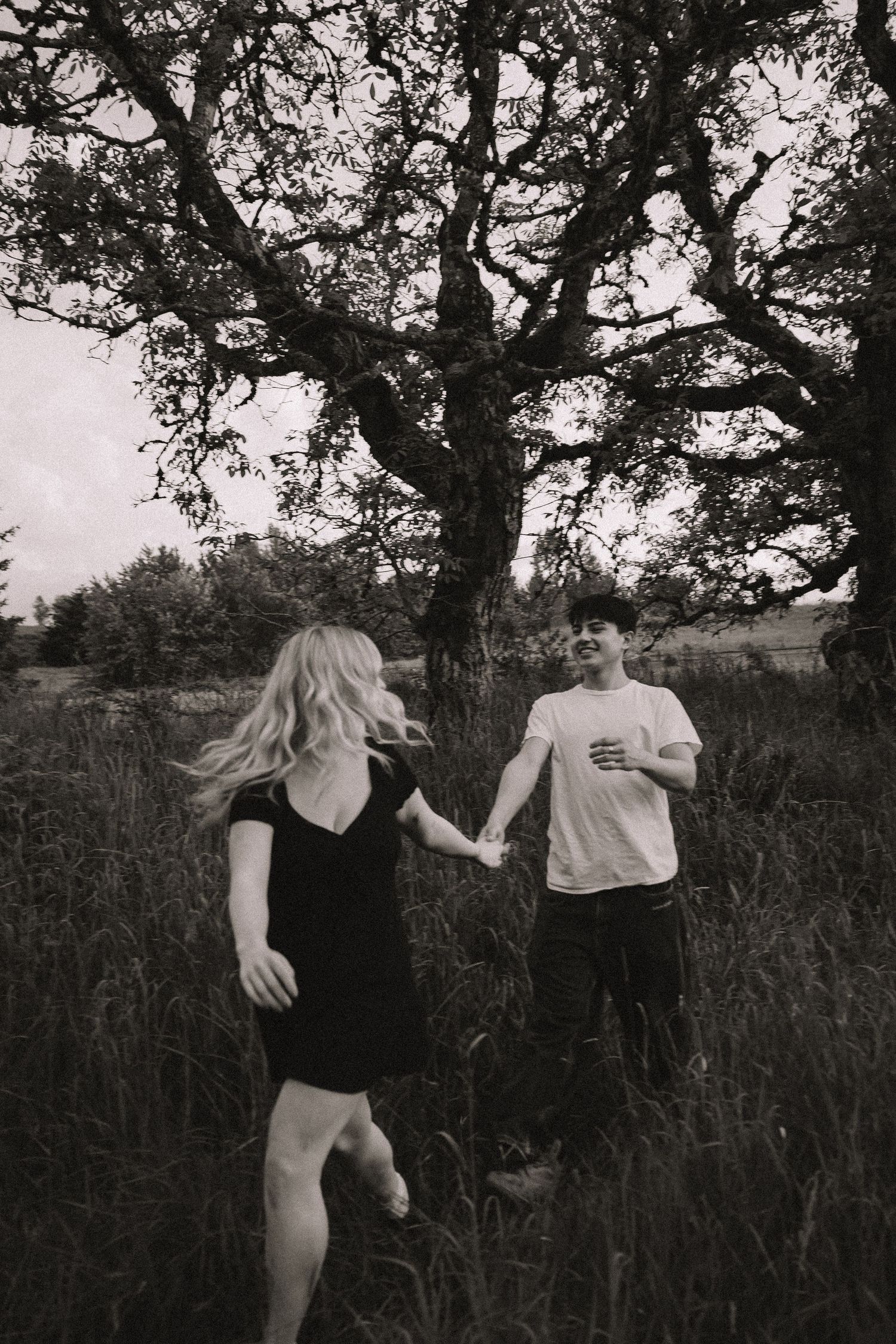 Black and white photo of a couple holding hands and running through a grassy field with a large tree behind them.