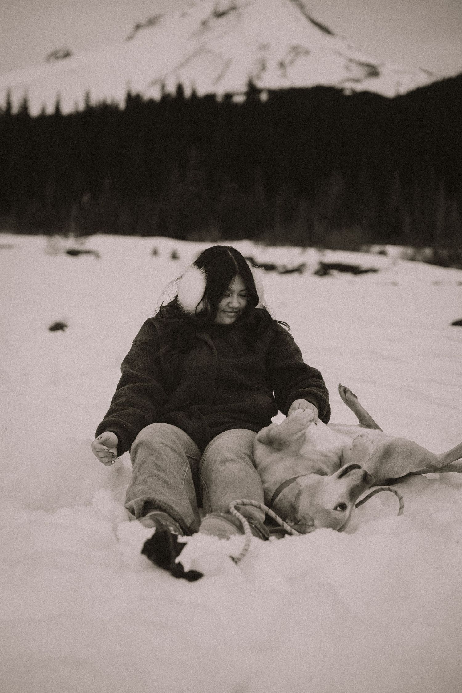Black and white photo of woman sitting in snow with a dog near a snow-covered mountain.