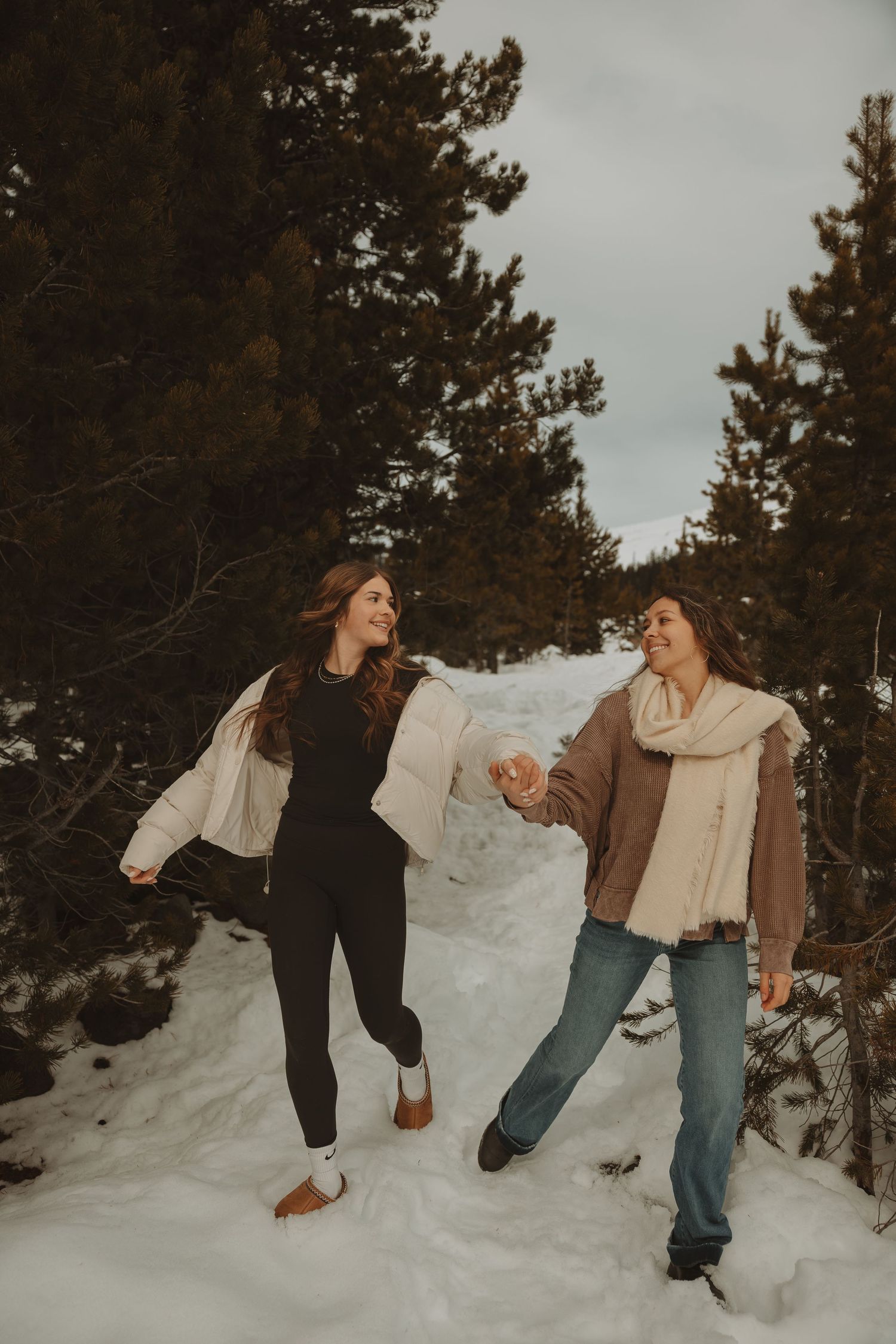 Two women laughing and holding hands while walking through a snowy pine forest in winter outfits.