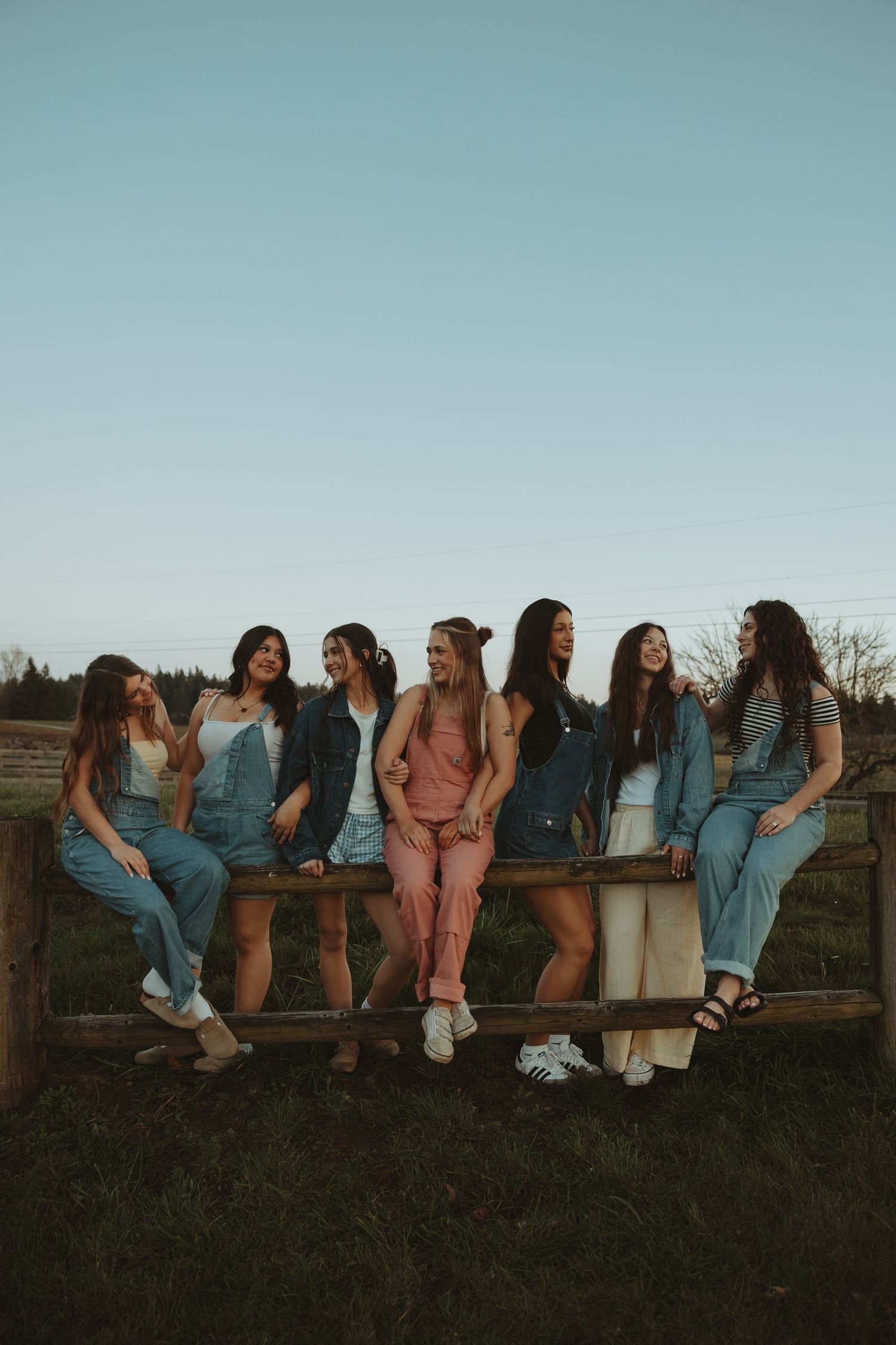 Group of seven women in casual denim outfits laughing and leaning on a rustic wooden fence at dusk.