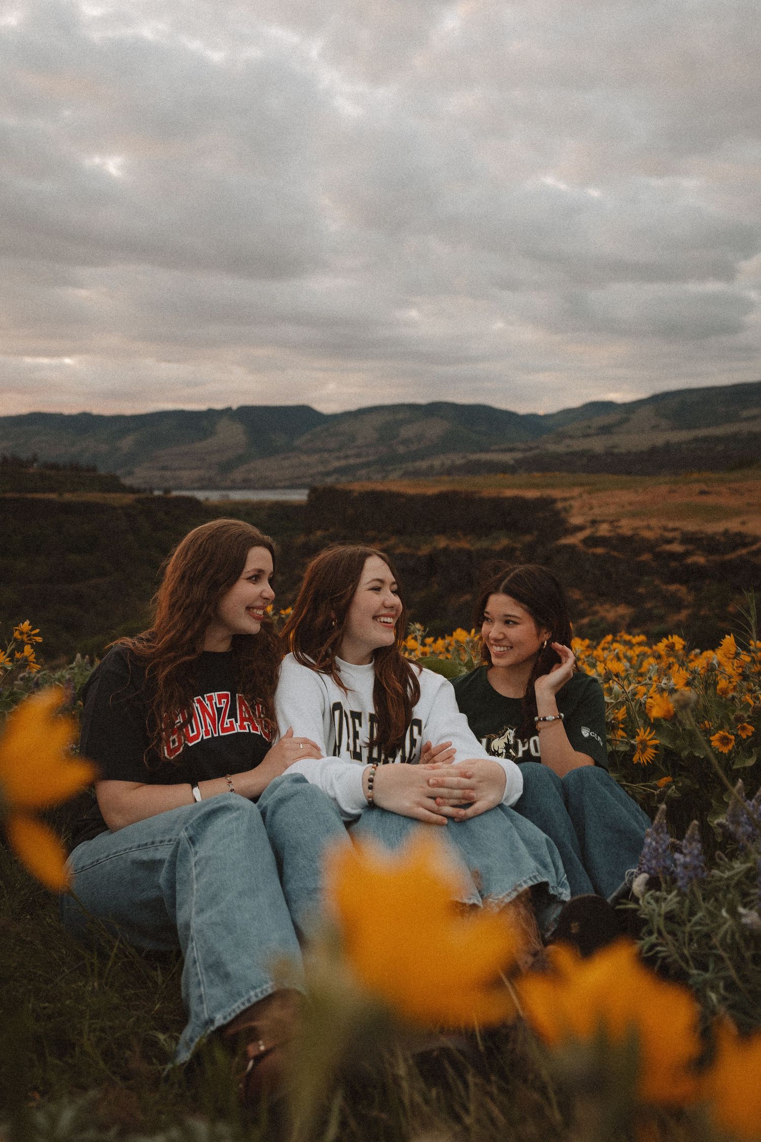 Three happy women laughing together in a golden wildflower meadow under cloudy skies.