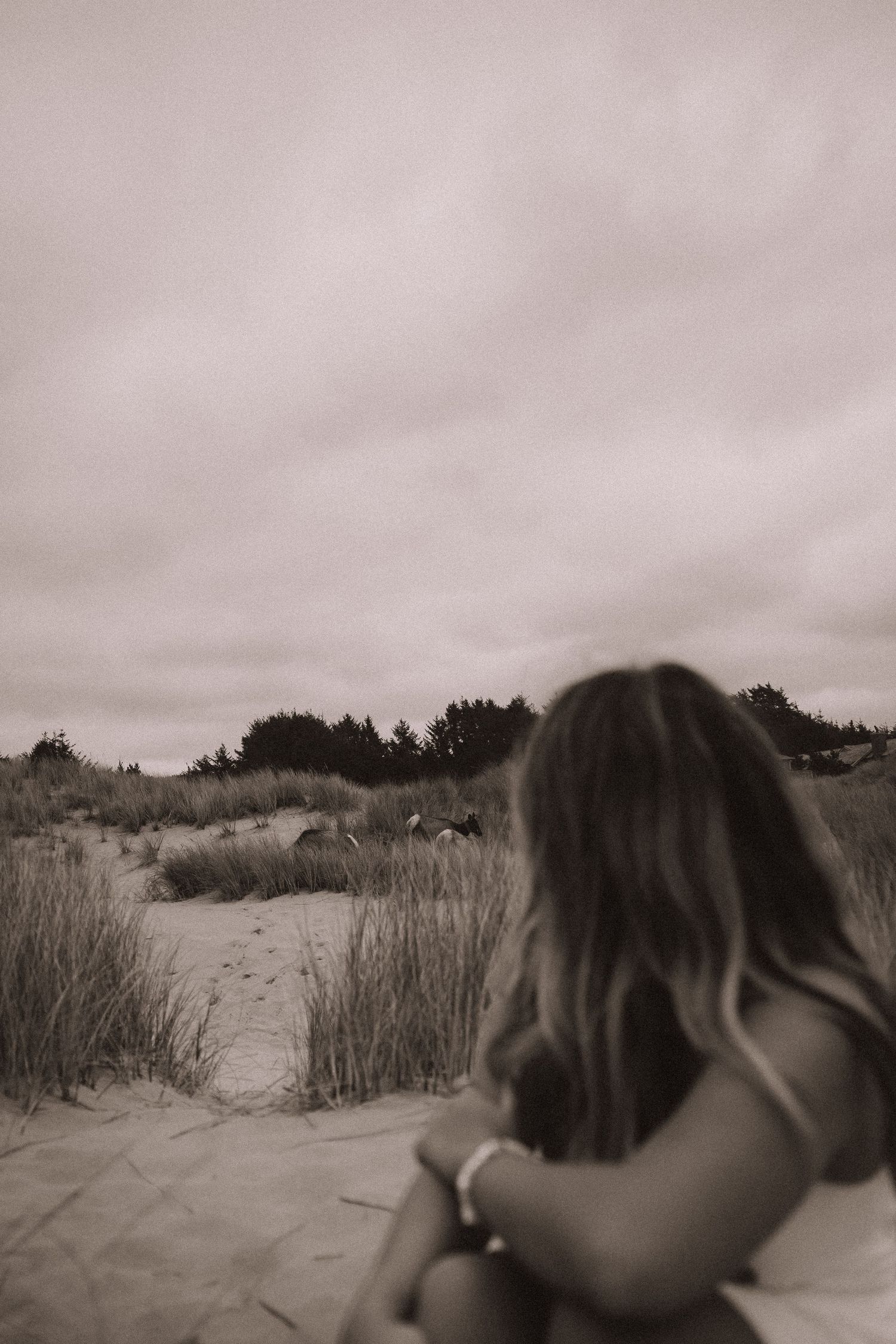 Black and white rear view of woman sitting on sand dunes with coastal grass and overcast sky backdrop.