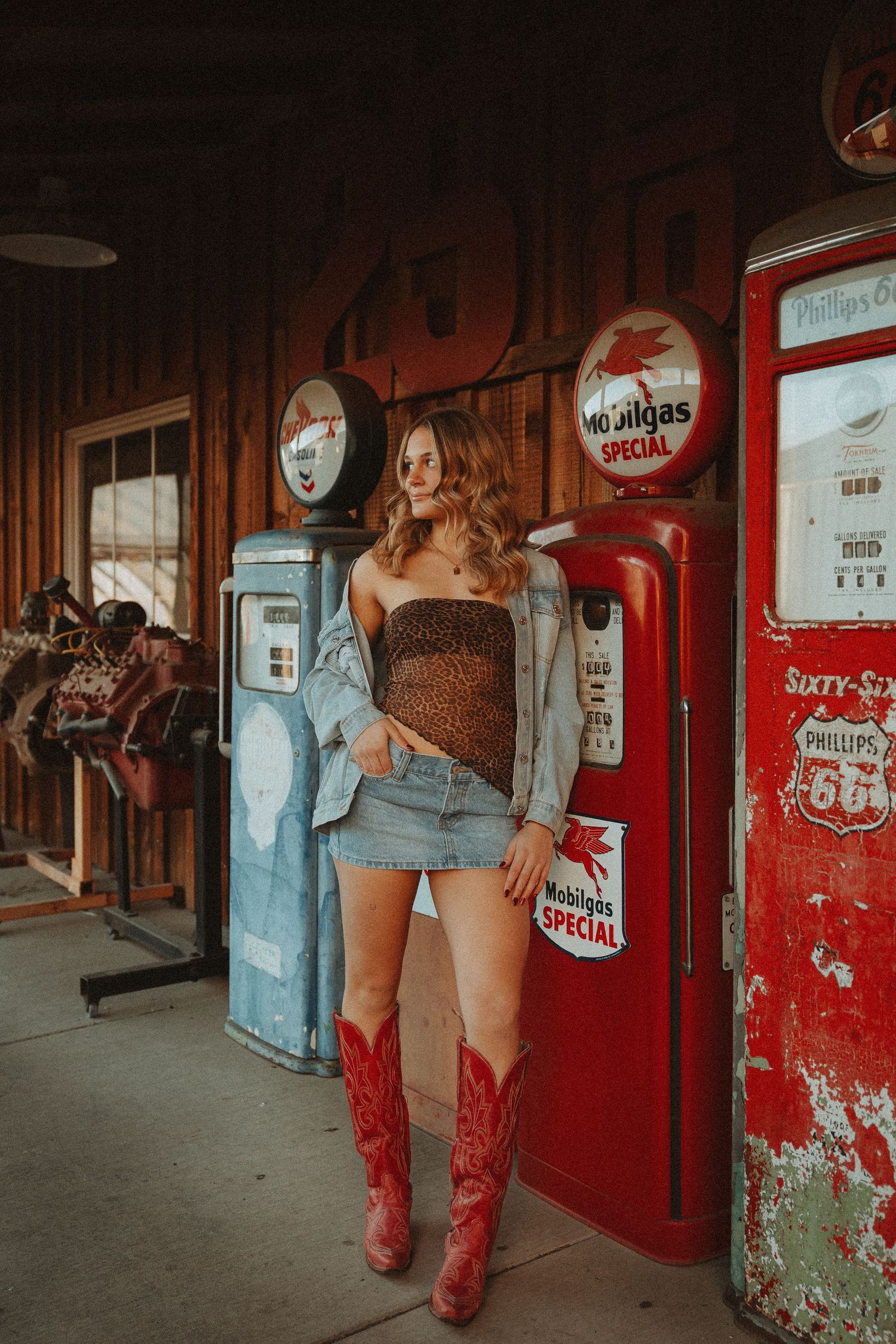 Woman in cowboy boots and denim skirt posing at vintage Phillips 66 gas station with retro Mobilgas pumps.