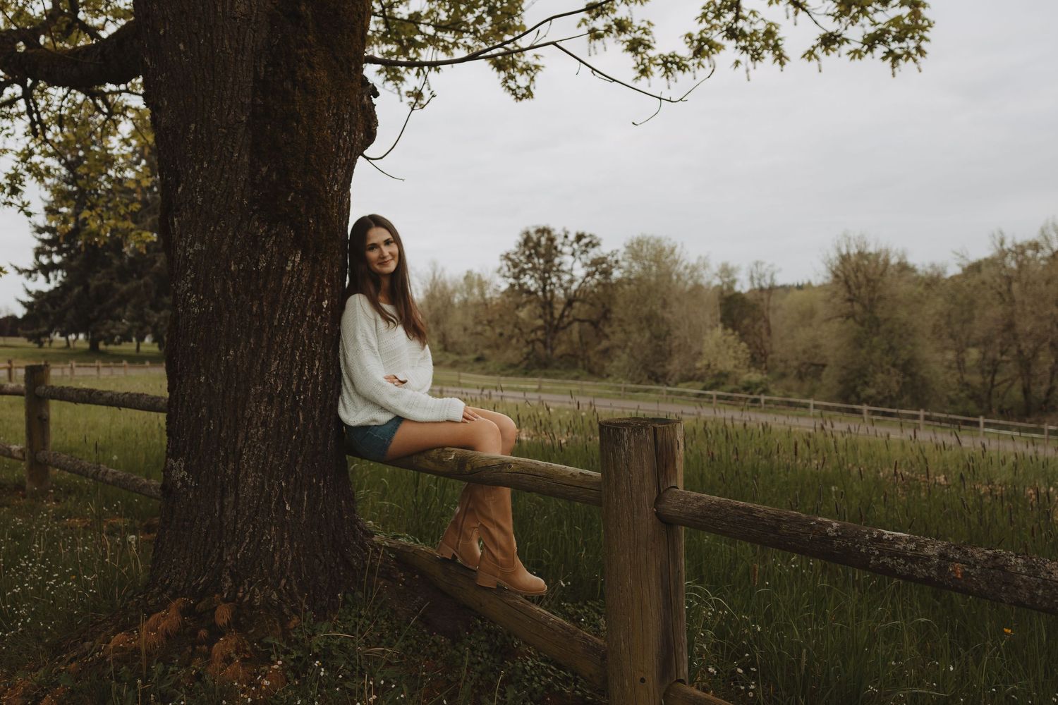 Young woman in white top and boots sitting on wooden fence beside a tree in a lush green countryside.