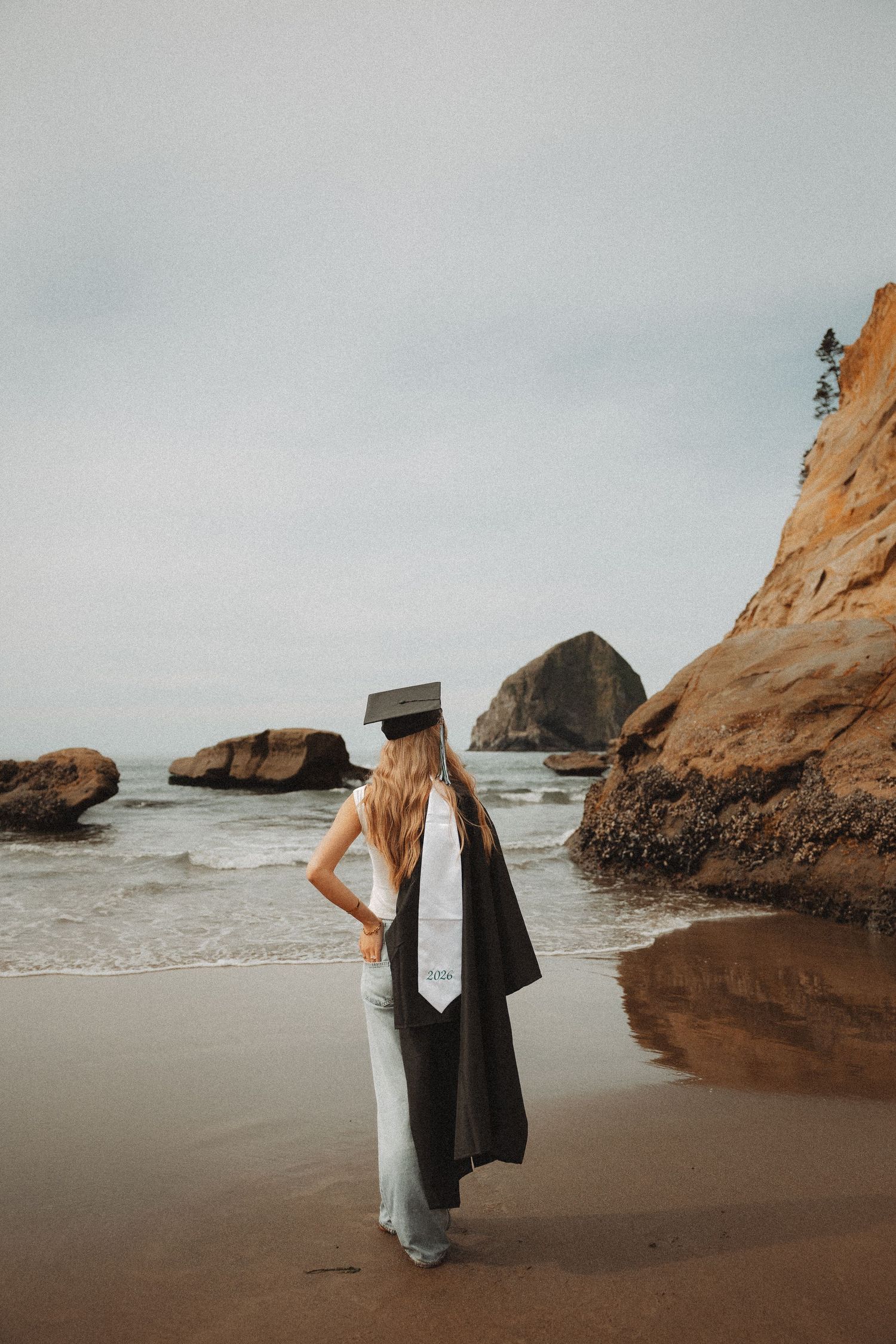Graduate in cap and gown stands on sandy beach gazing at ocean waves and rocky cliffs on overcast day.