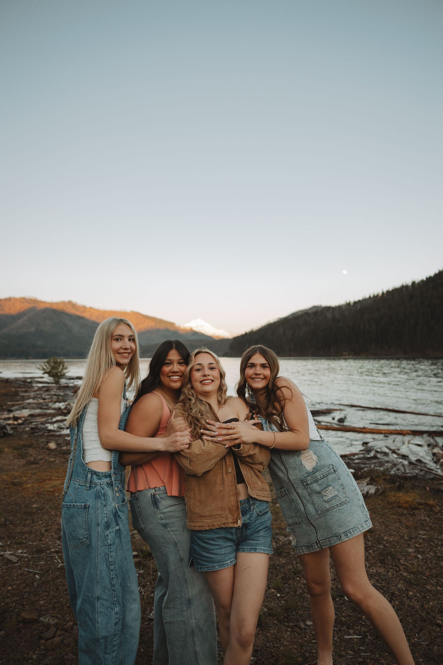 Four smiling young women hugging together by a mountain lake at sunset.