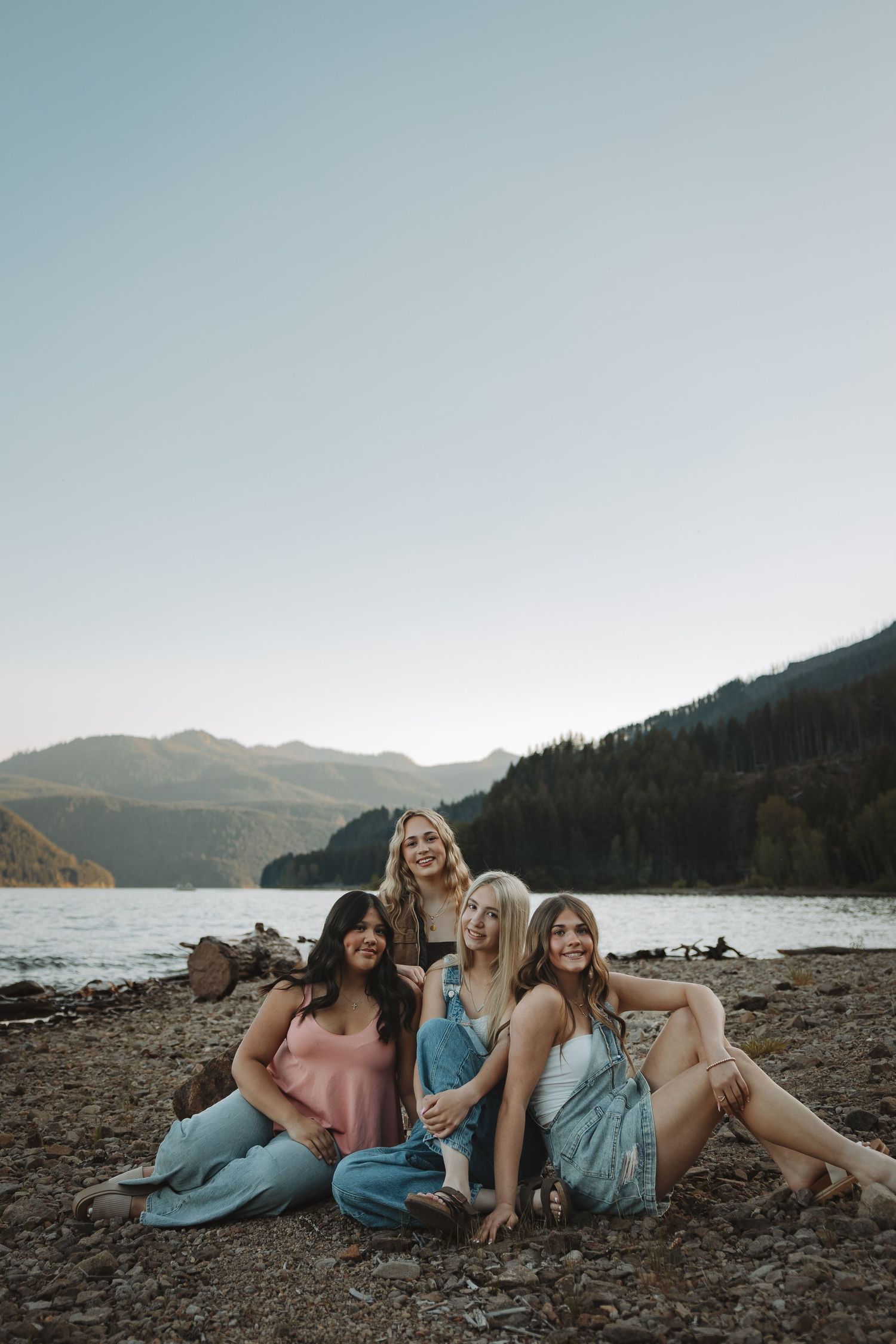 Four smiling young women posing together on a rocky lakeshore with mountain scenery at sunset.