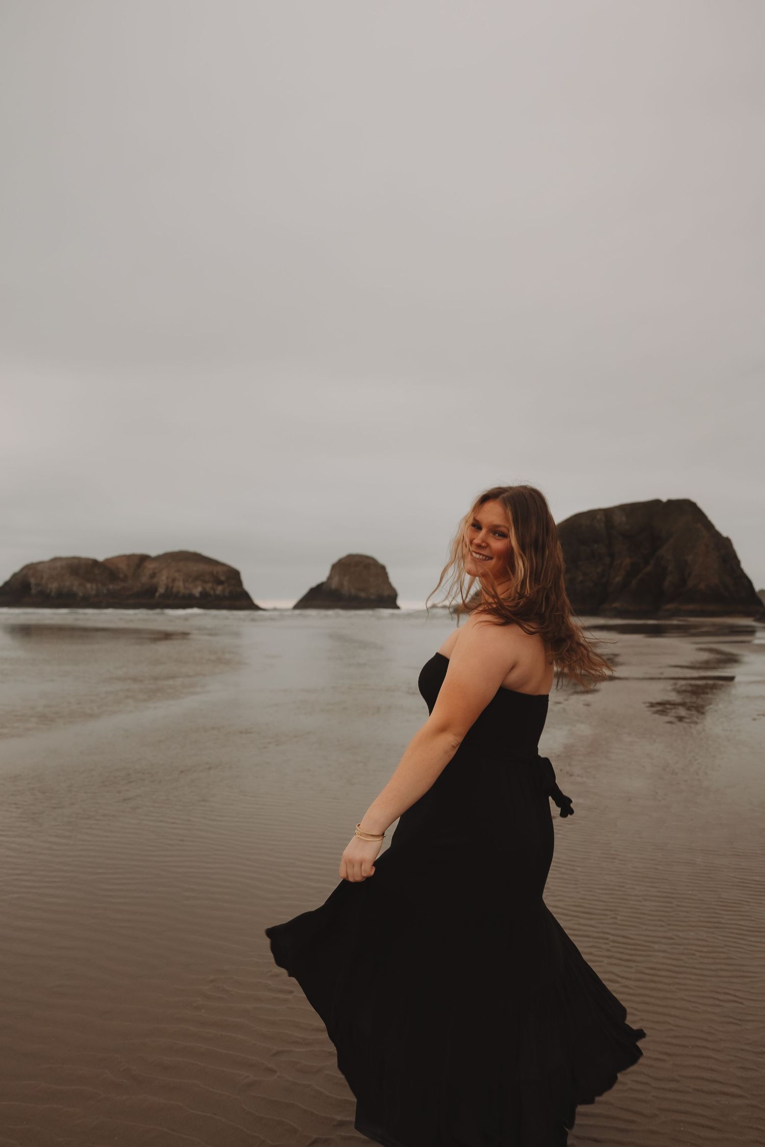 A figure in a flowing black dress twirls gracefully on a misty beach with rocky outcrops visible in the background.