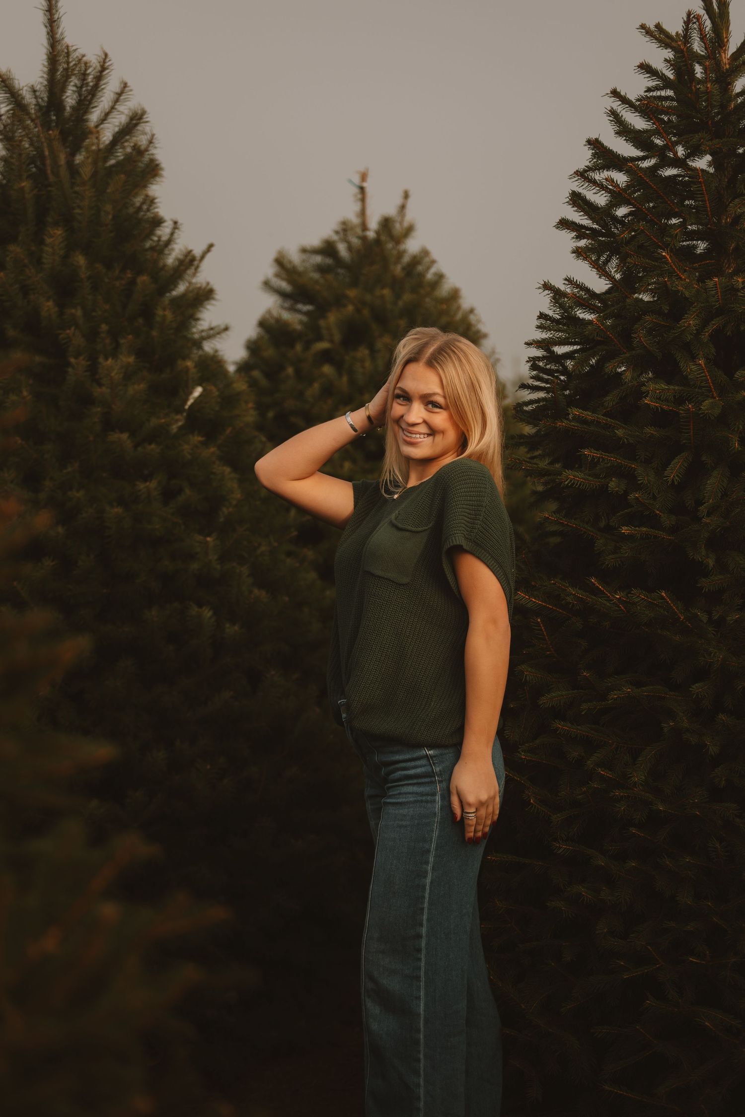 Person in green top and denim poses cheerfully against backdrop of evergreen trees.