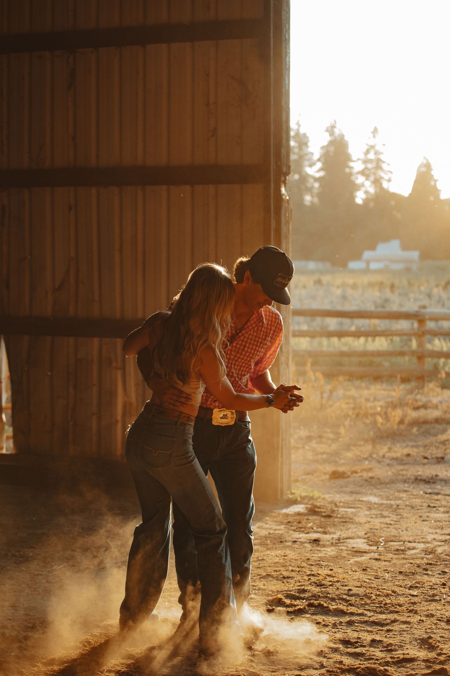 Couple dancing together in golden sunset light on a dusty ranch beside a wooden barn.
