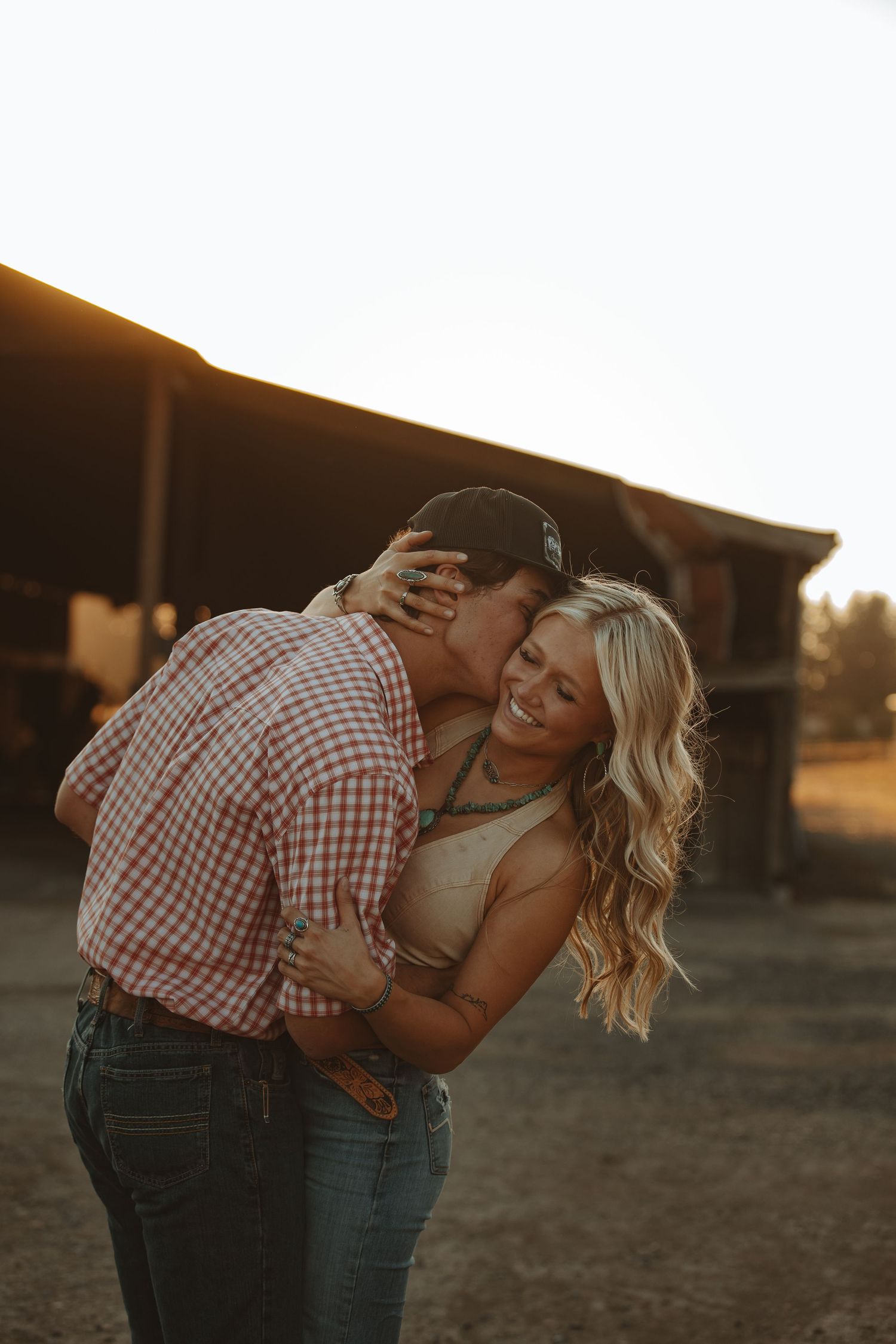 Man in plaid shirt embracing smiling blonde woman at sunset near a rustic barn on a farm.