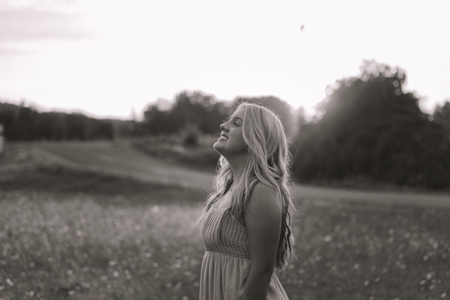Black and white photo of a young woman with long blonde hair standing in a field, head tilted back, smiling.