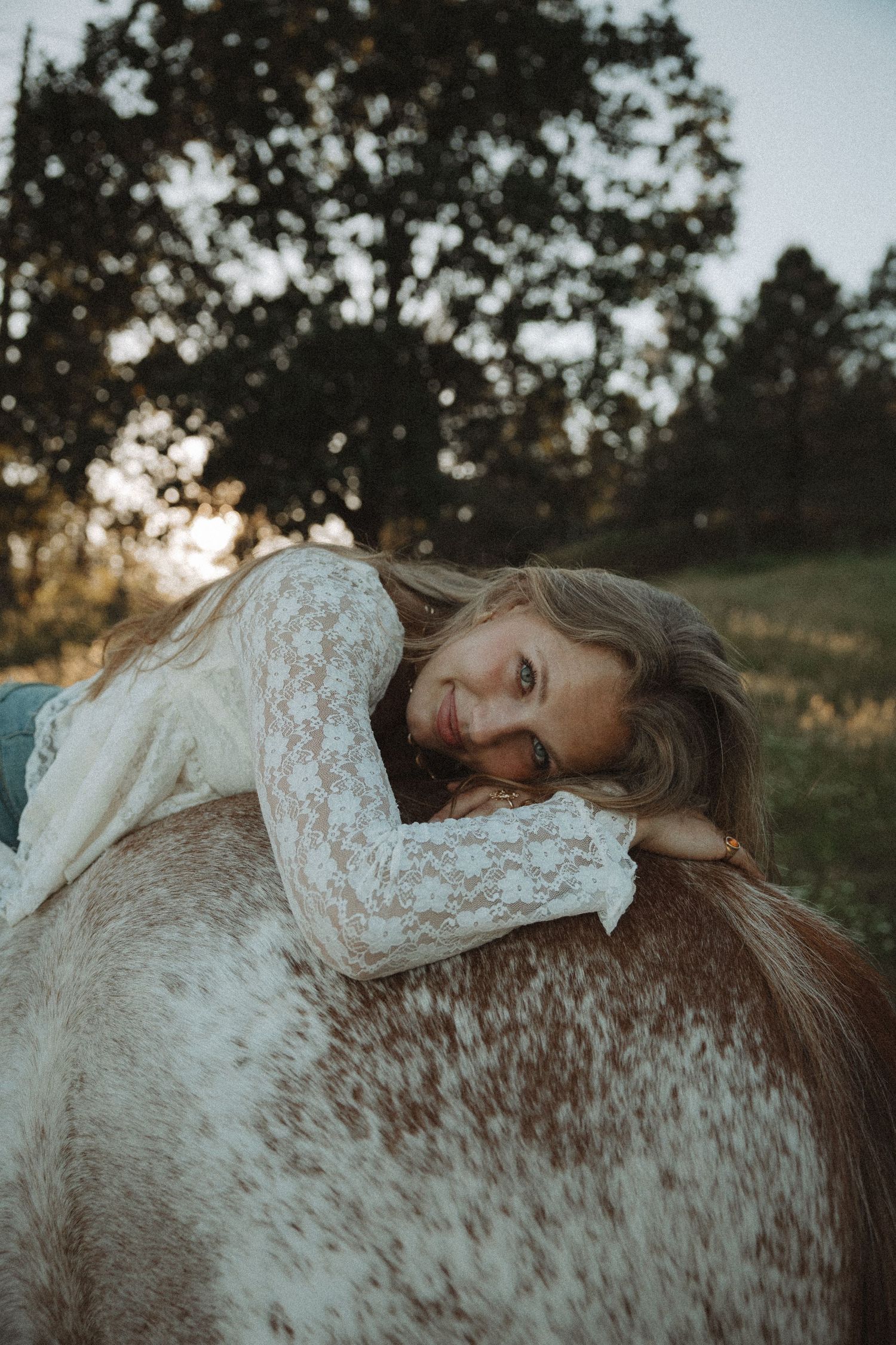 Young woman in white lace top resting on a horse in a sunlit field with trees in background.