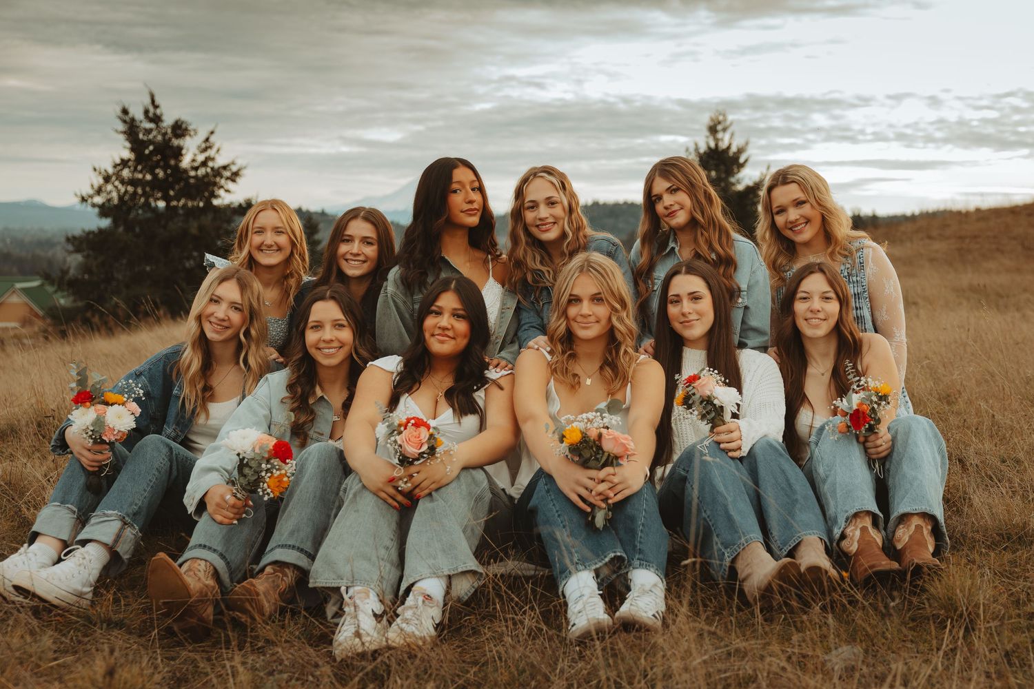 A group of friends in matching denim and white outfits pose together on a grassy hillside at sunset.