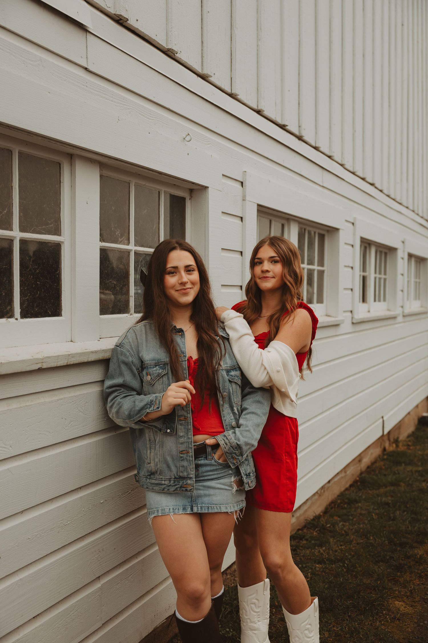 Two women in denim jacket and red top pose casually against white building exterior.
