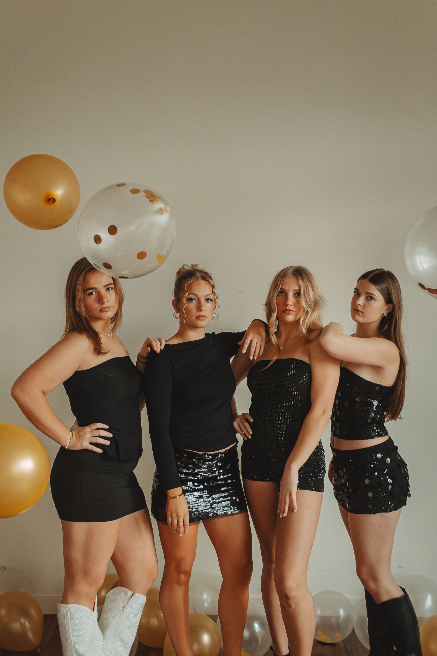 A group poses playfully in black outfits and sequin shorts with gold balloons in a party setting.