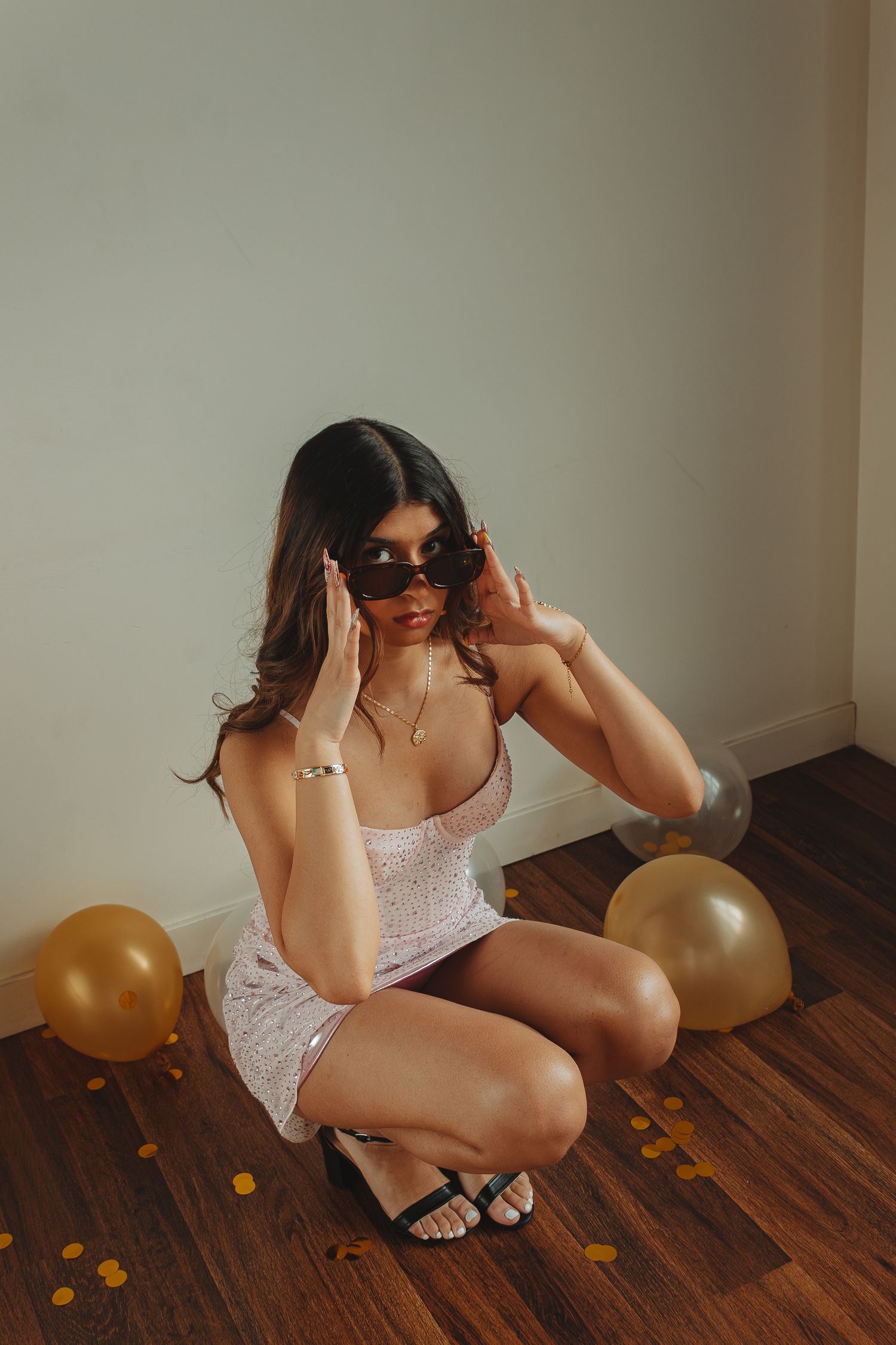 Person in white dress and sunglasses posing with gold balloons on wooden floor.