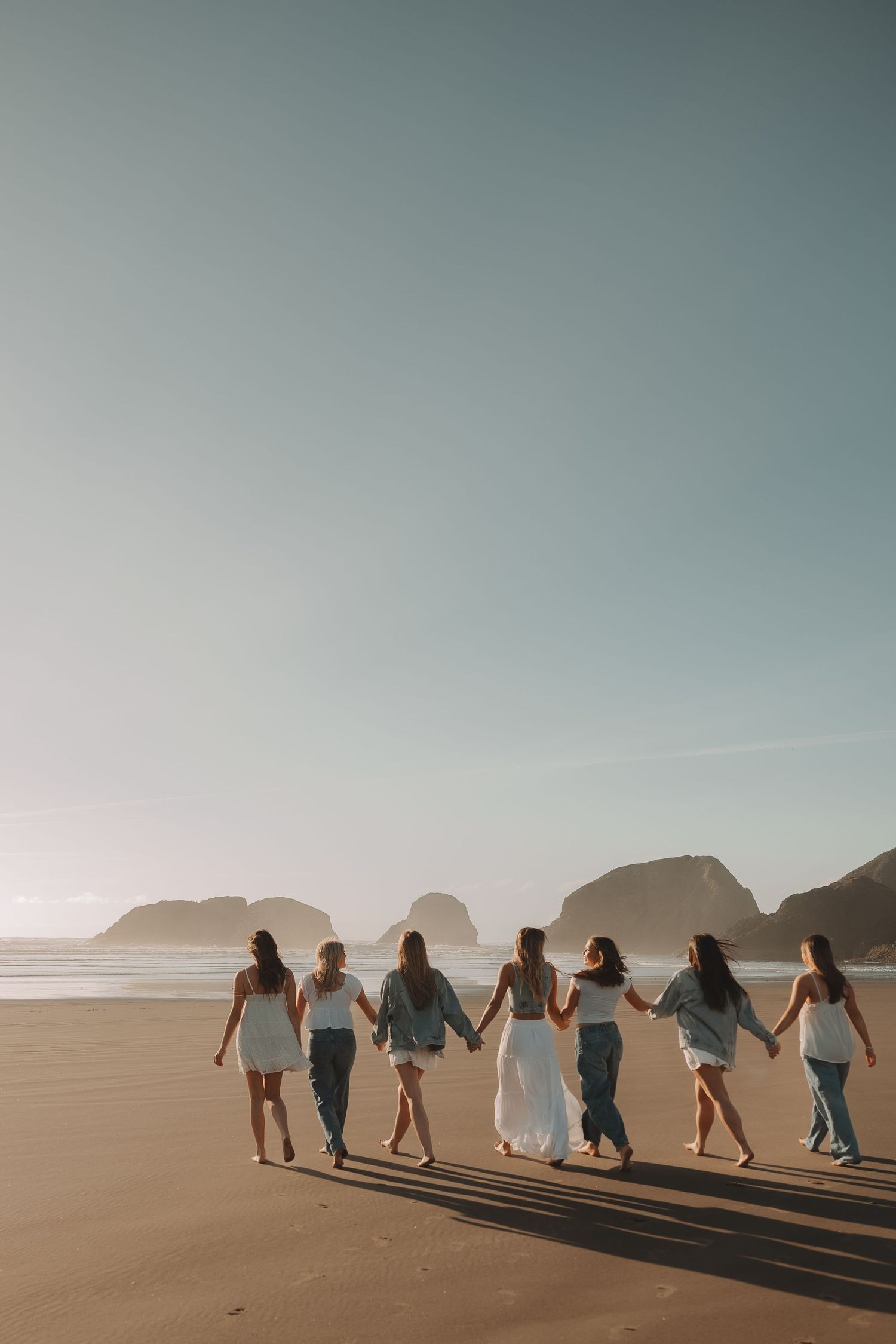 Six women in white and blue outfits hold hands walking along a scenic beach at golden hour.