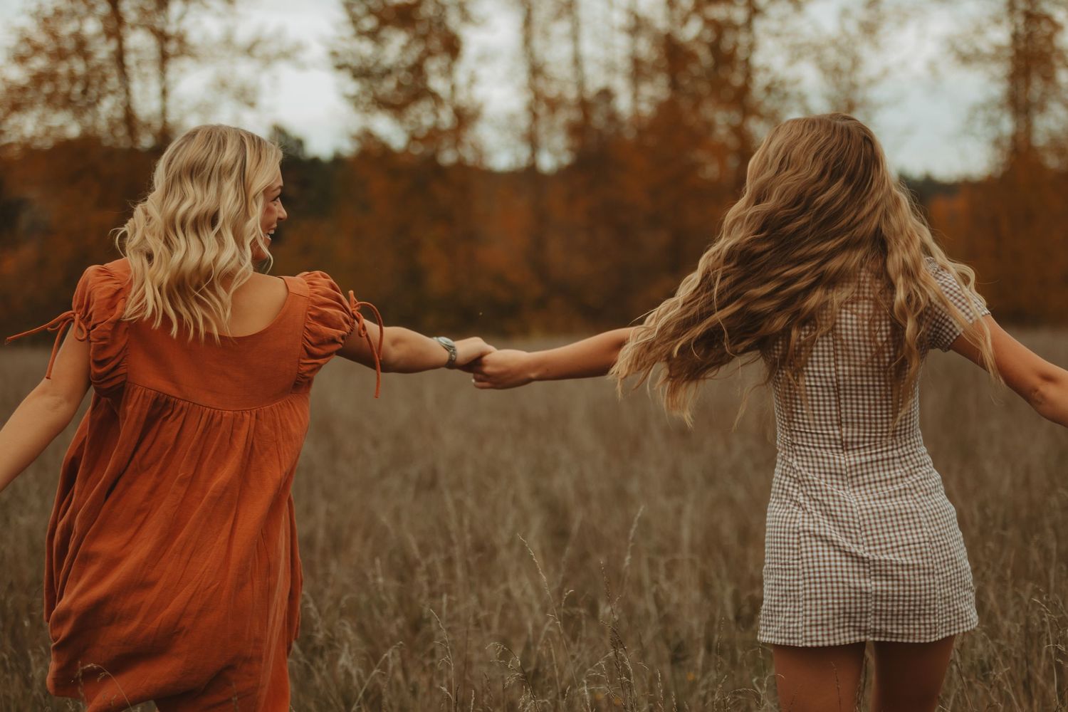 Two people in summer dresses hold hands while standing in a golden field during sunset.
