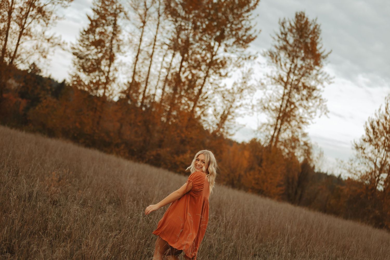 Person in orange sweater stands in golden field with autumn trees in background.