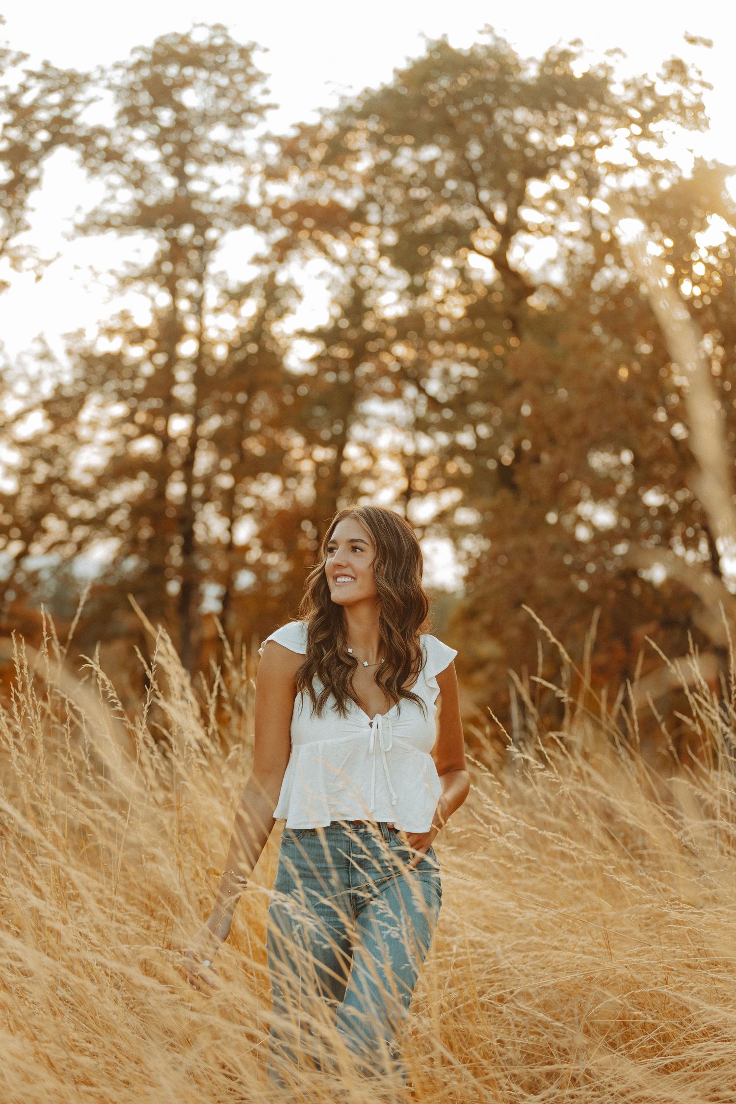 Woman in white top sitting in golden field of tall grass during sunset with warm autumn lighting.