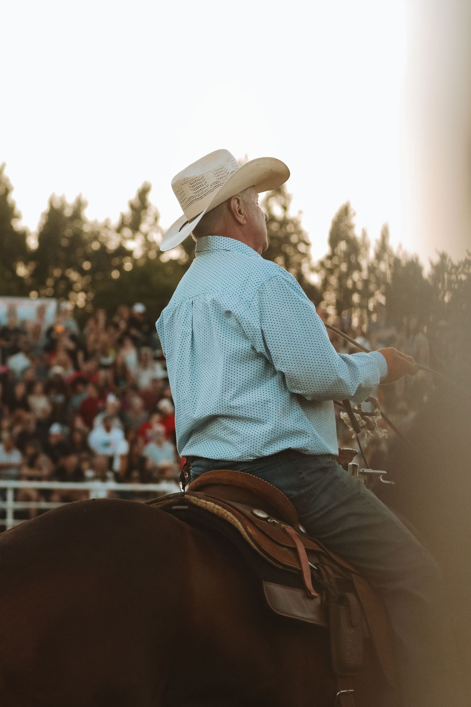 Cowboy in light blue shirt and hat sits on horseback at sunset during rodeo event.
