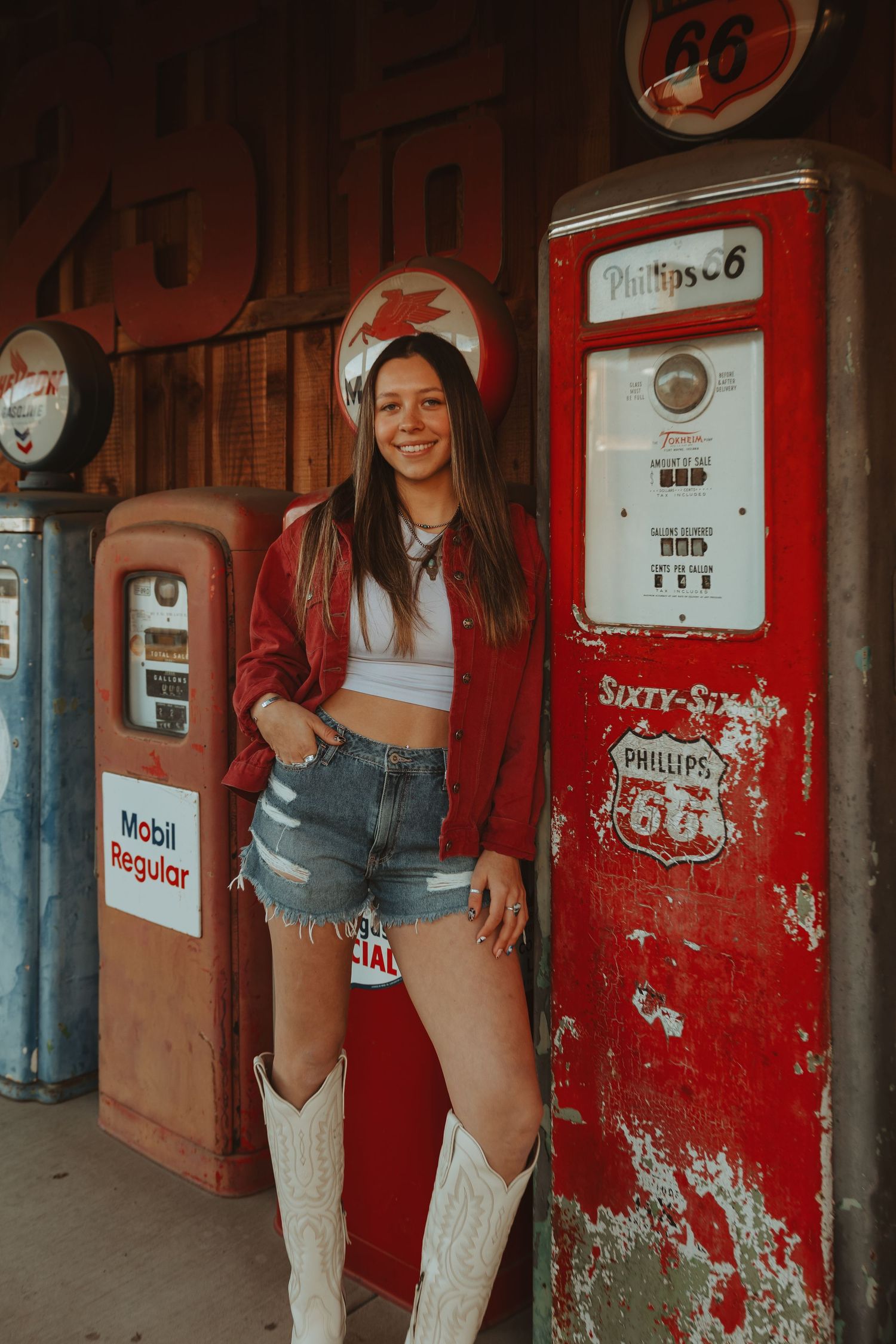 Young woman in red jacket, denim shorts, and white cowboy boots posing by vintage Phillips 66 gas pumps.