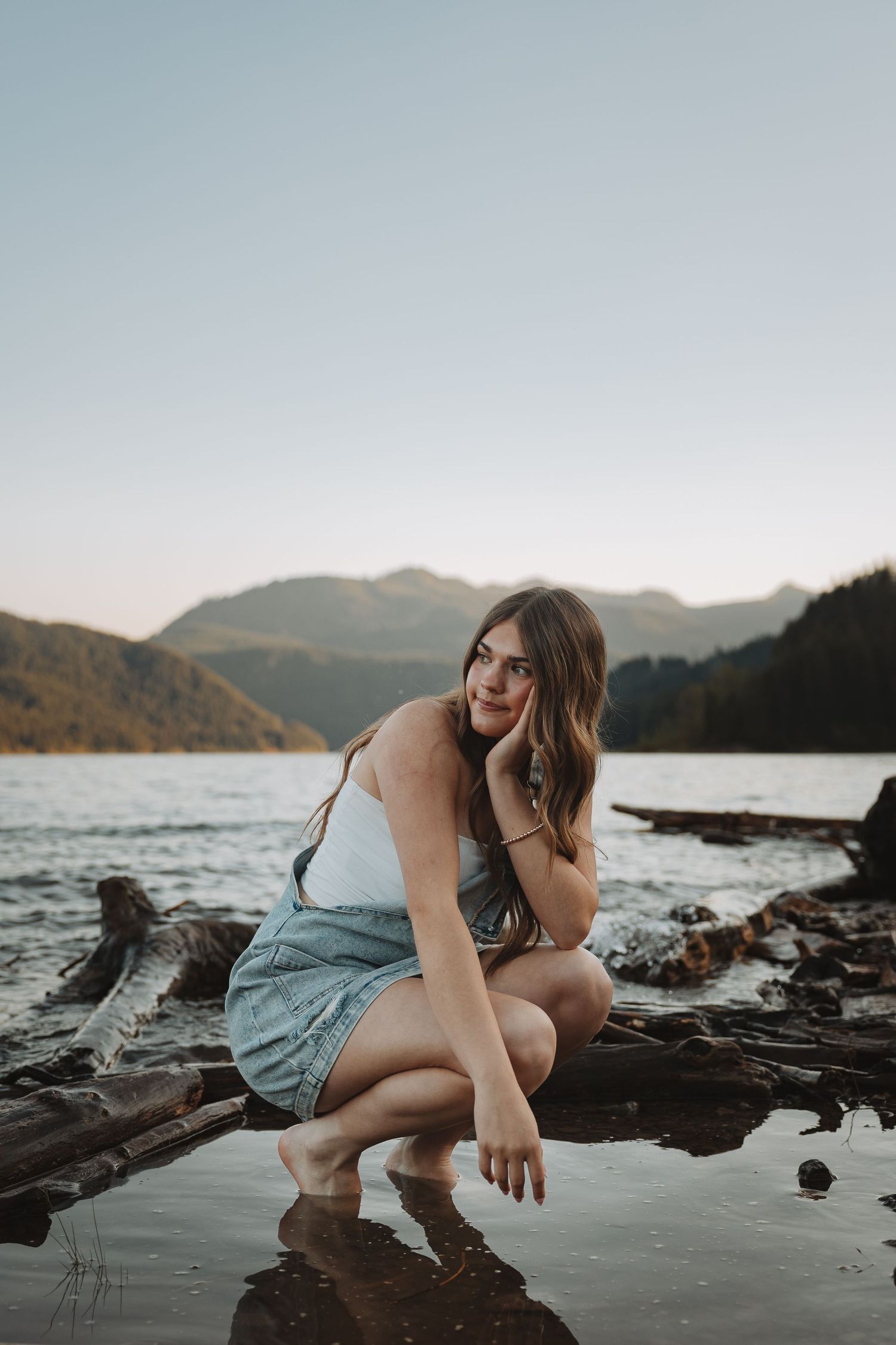Young woman in denim overalls crouching on driftwood by a mountain lake at sunset.