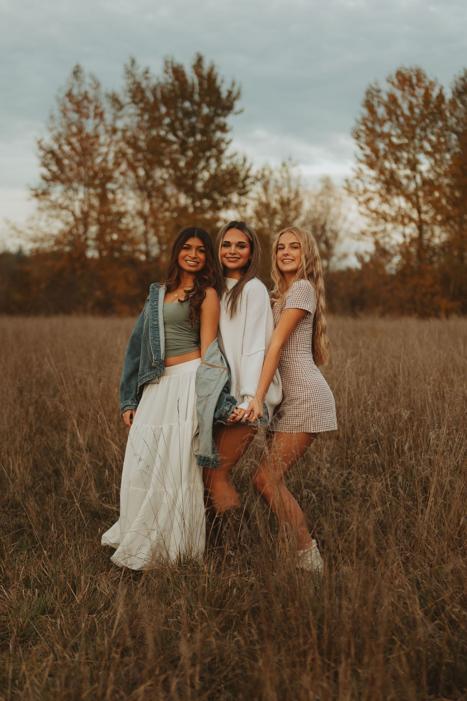 Three people pose together in casual attire against a backdrop of tall grass and autumn trees.