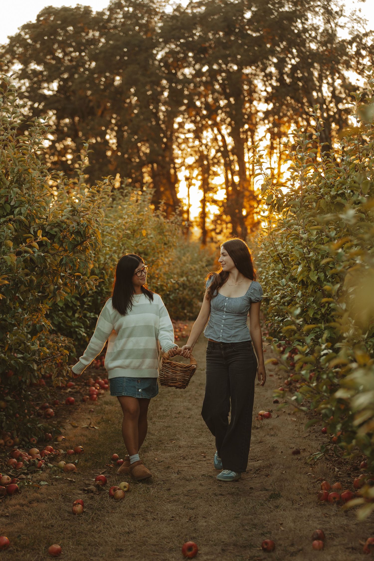 Two people walking together through rows of apple trees at sunset while carrying baskets.