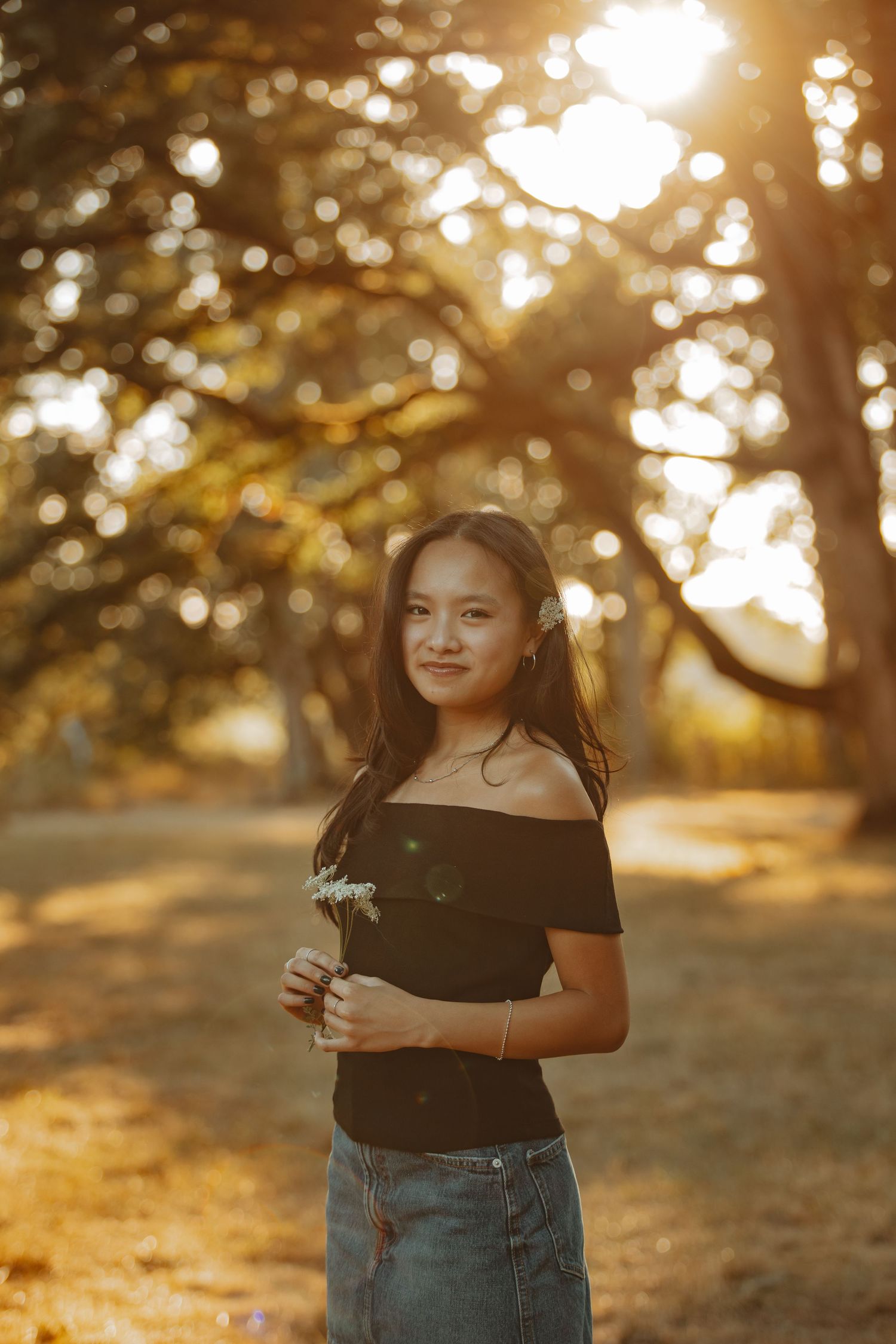A young person stands in a sunlit forest holding a flower, wearing a black top and denim skirt during golden hour.