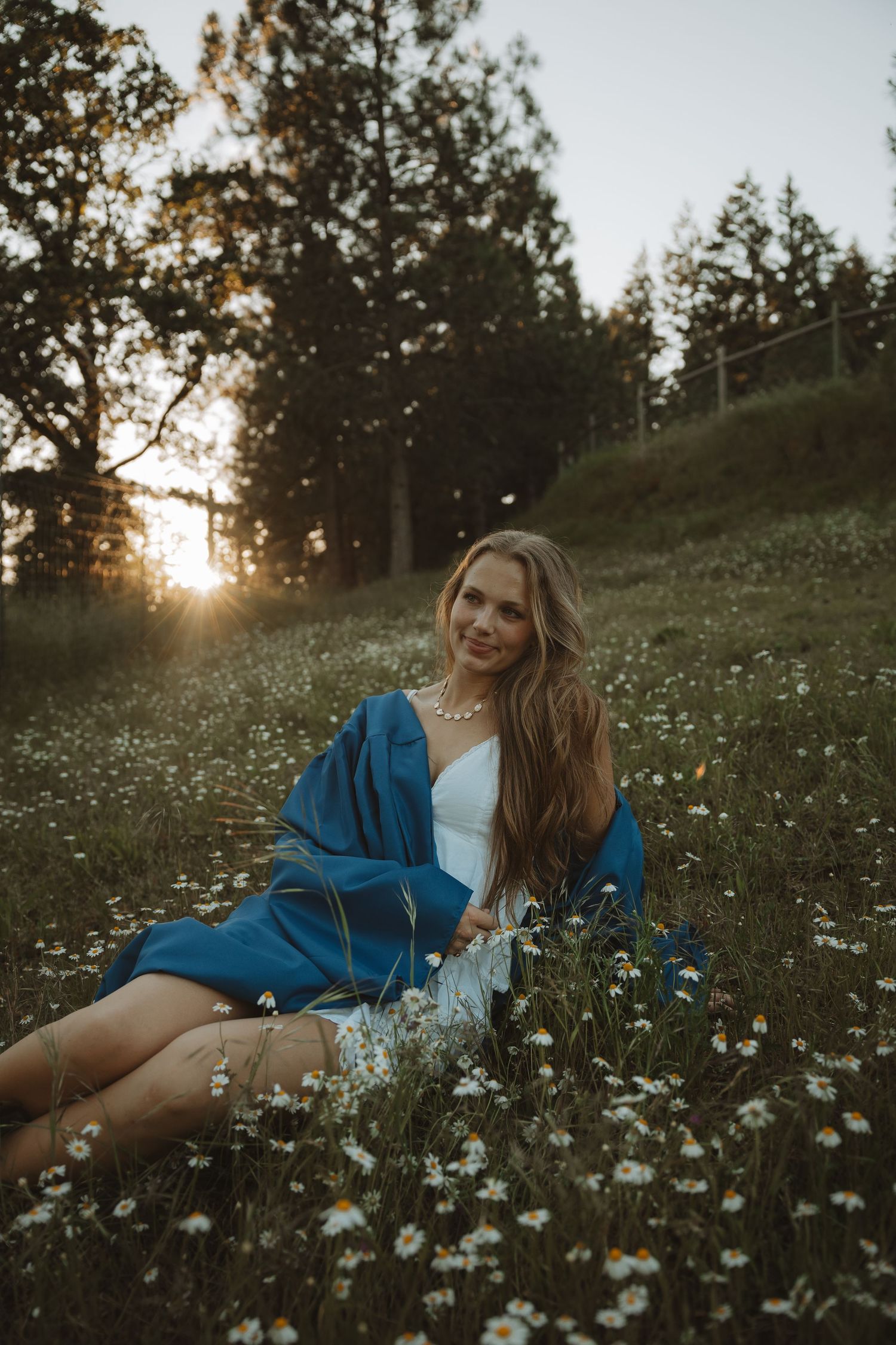 Young woman in blue robe sitting in wildflower meadow at golden sunset, smiling at camera.
