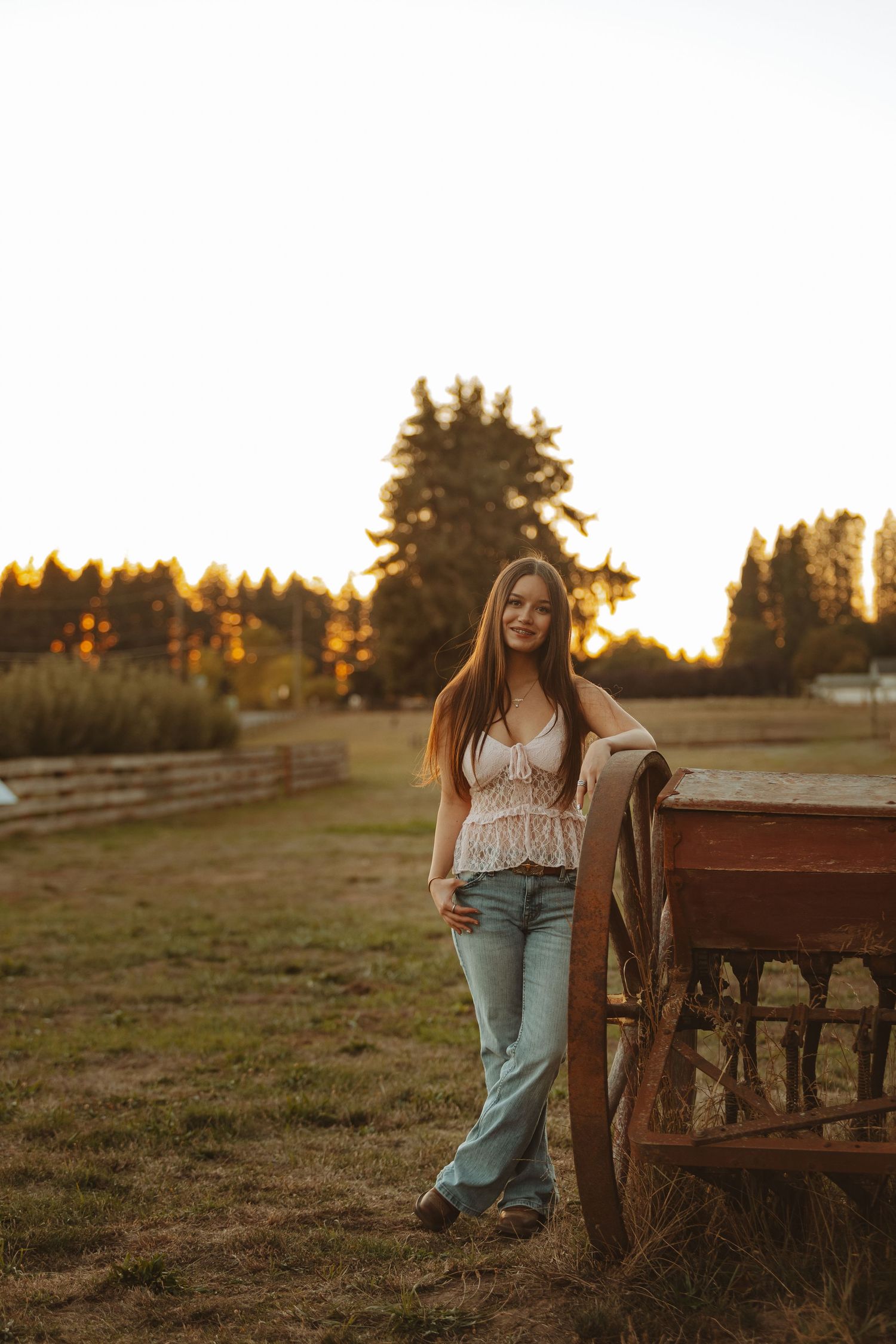 Young woman in lace top and jeans leaning on rustic wooden wagon at golden hour on a farm.