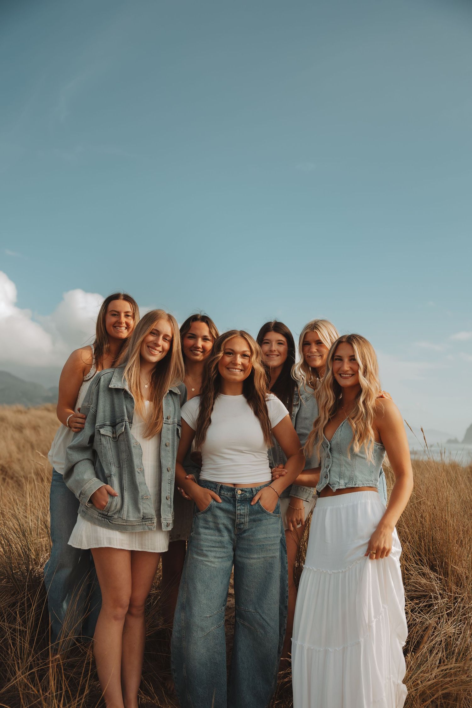 Seven smiling young women pose together in a golden grass field under a blue sky.