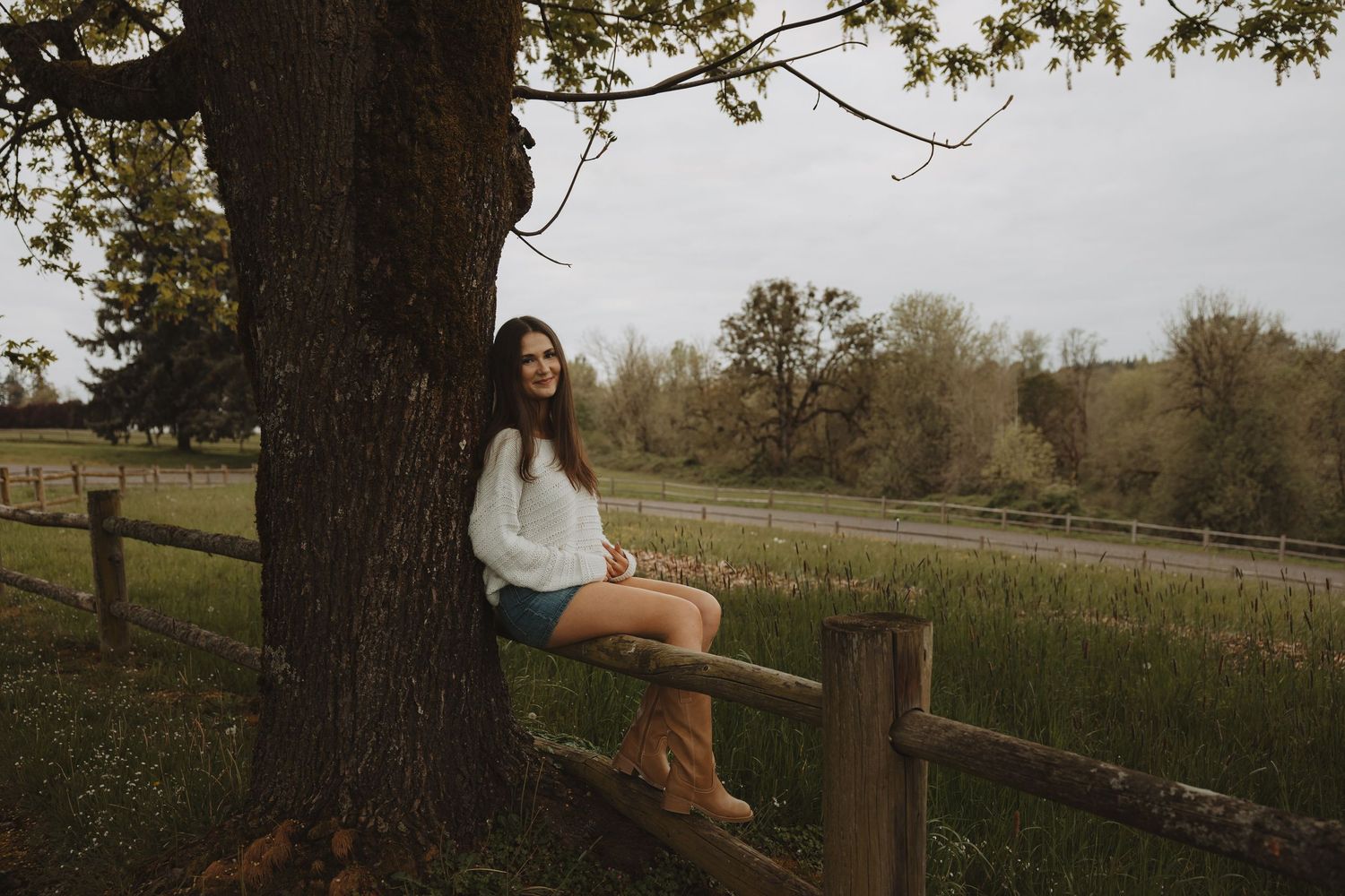 Young woman in white sweater and cowboy boots sitting on a wooden fence beside a large tree in a rural setting.
