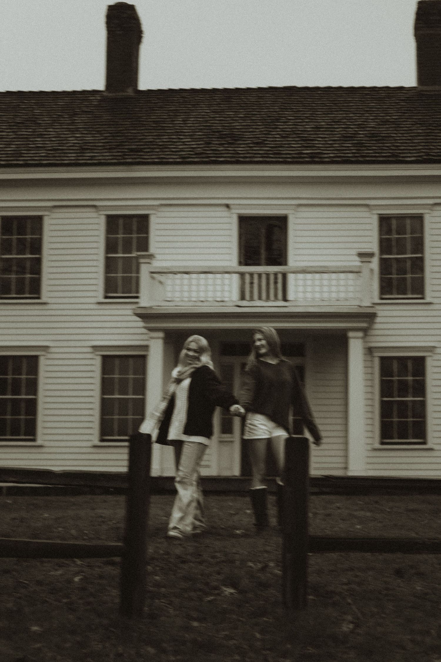 A black and white photo of two people standing in front of a historic white colonial house with a balcony.